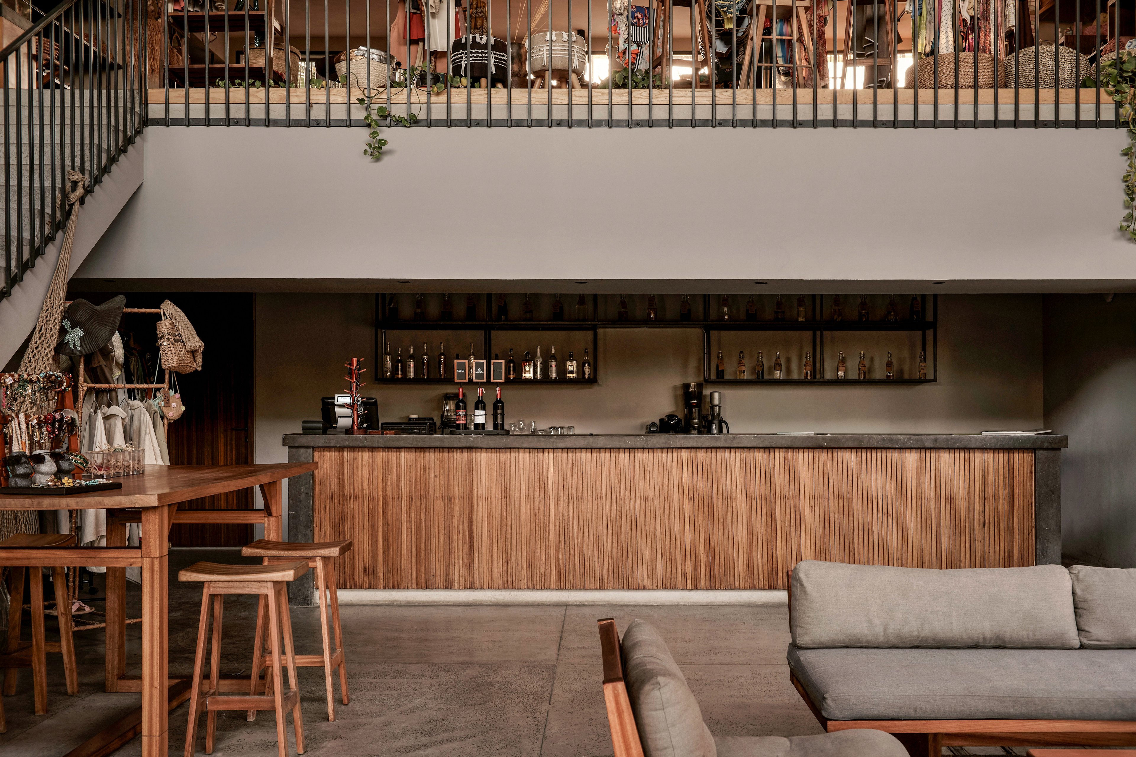 A contemporary cafe featuring a minimalist design with wooden furniture and a sleek bar counter. The setting includes a cozy seating area with sofas and stools, complemented by a mezzanine level with additional decor. A person is visible behind the bar, adding a human element to the scene.