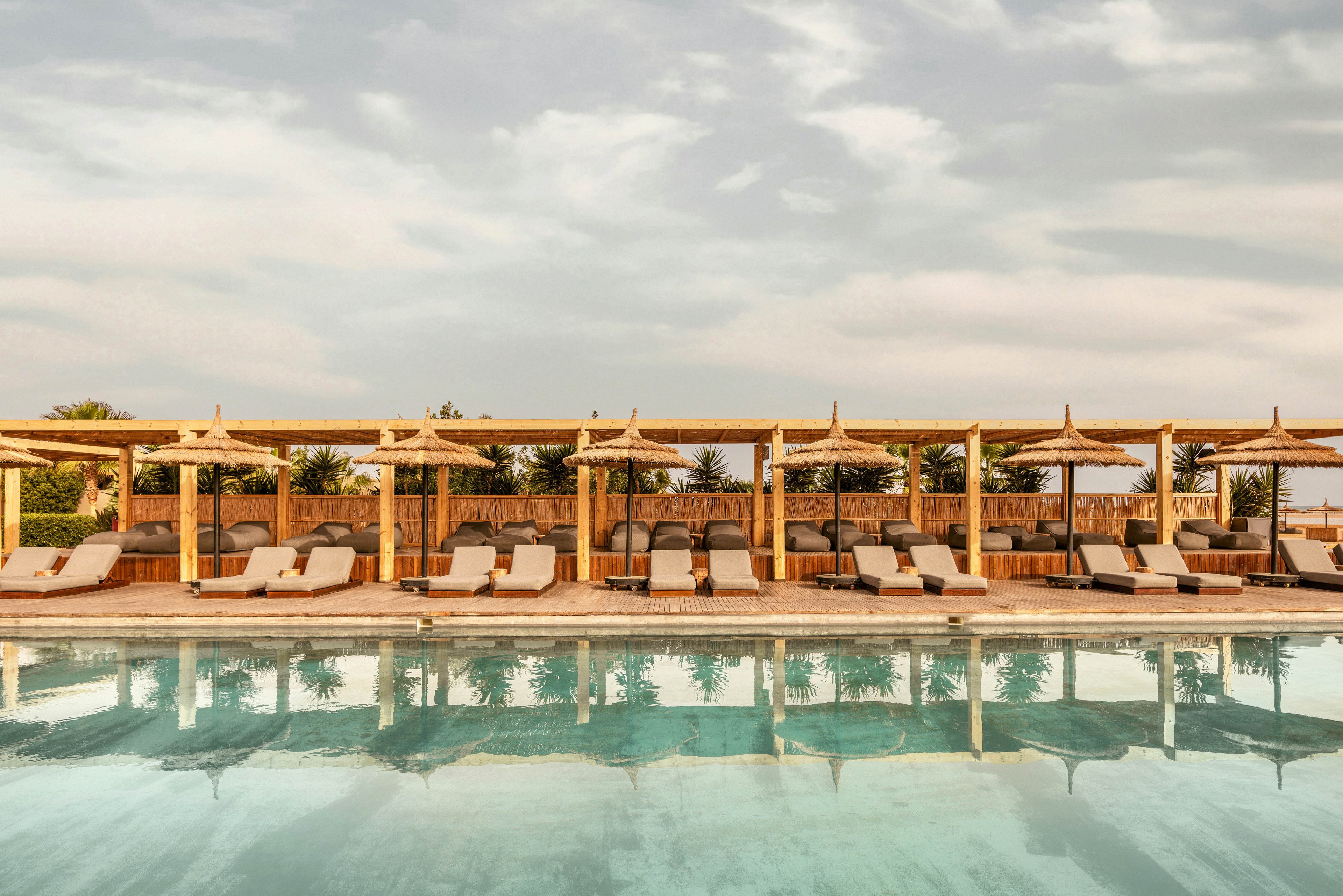 A serene outdoor pool area featuring wooden cabanas with straw roofs and cushioned lounge chairs. The pool reflects the clear sky, creating a tranquil atmosphere. Palm trees and greenery enhance the tropical resort vibe.