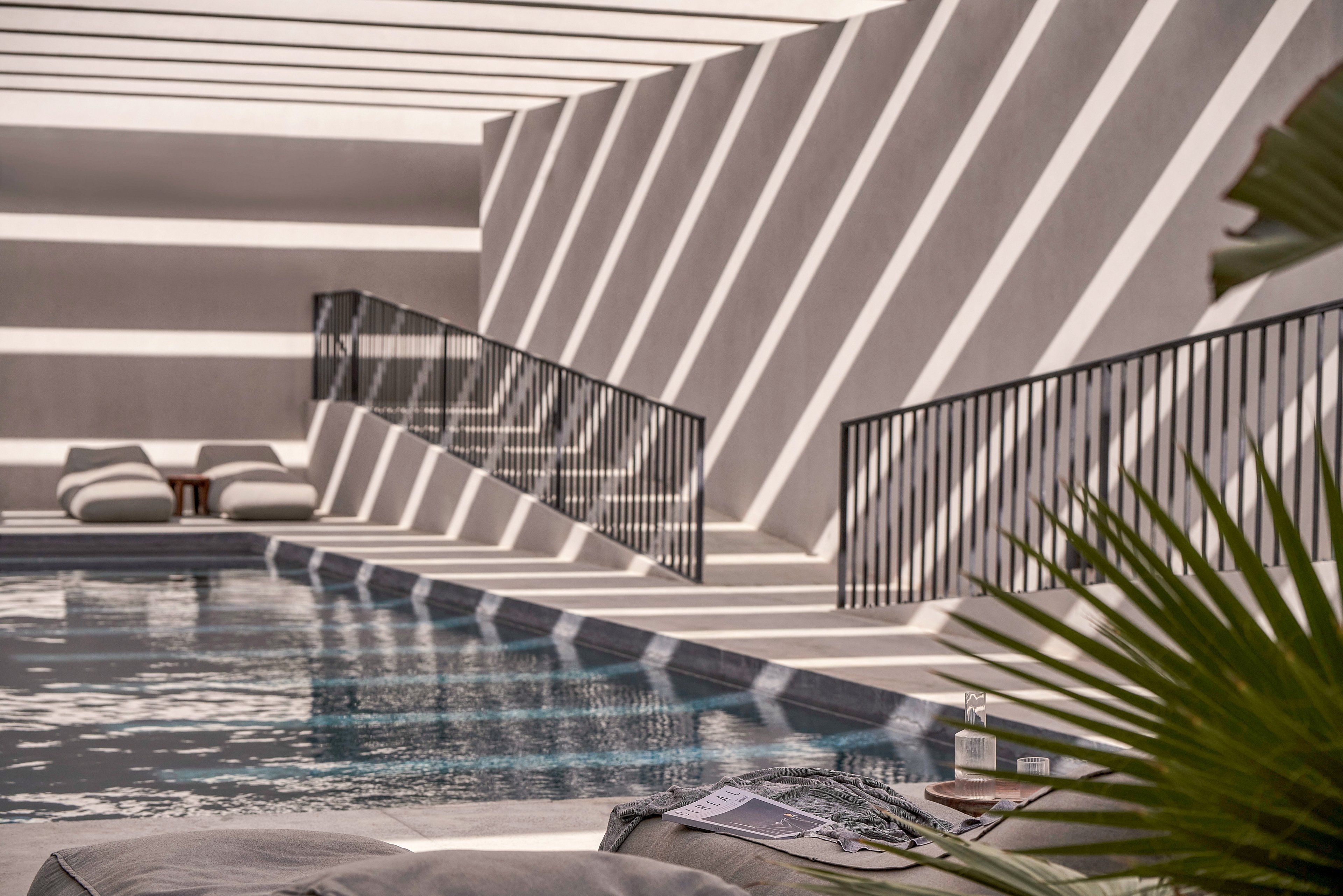 A serene poolside setting featuring modern architecture with bold geometric shadows. The space includes minimalist lounge chairs and a sleek railing design. The soft lighting and reflections on the water create a tranquil atmosphere. A small table with a bottle and glass adds a touch of detail to the scene.