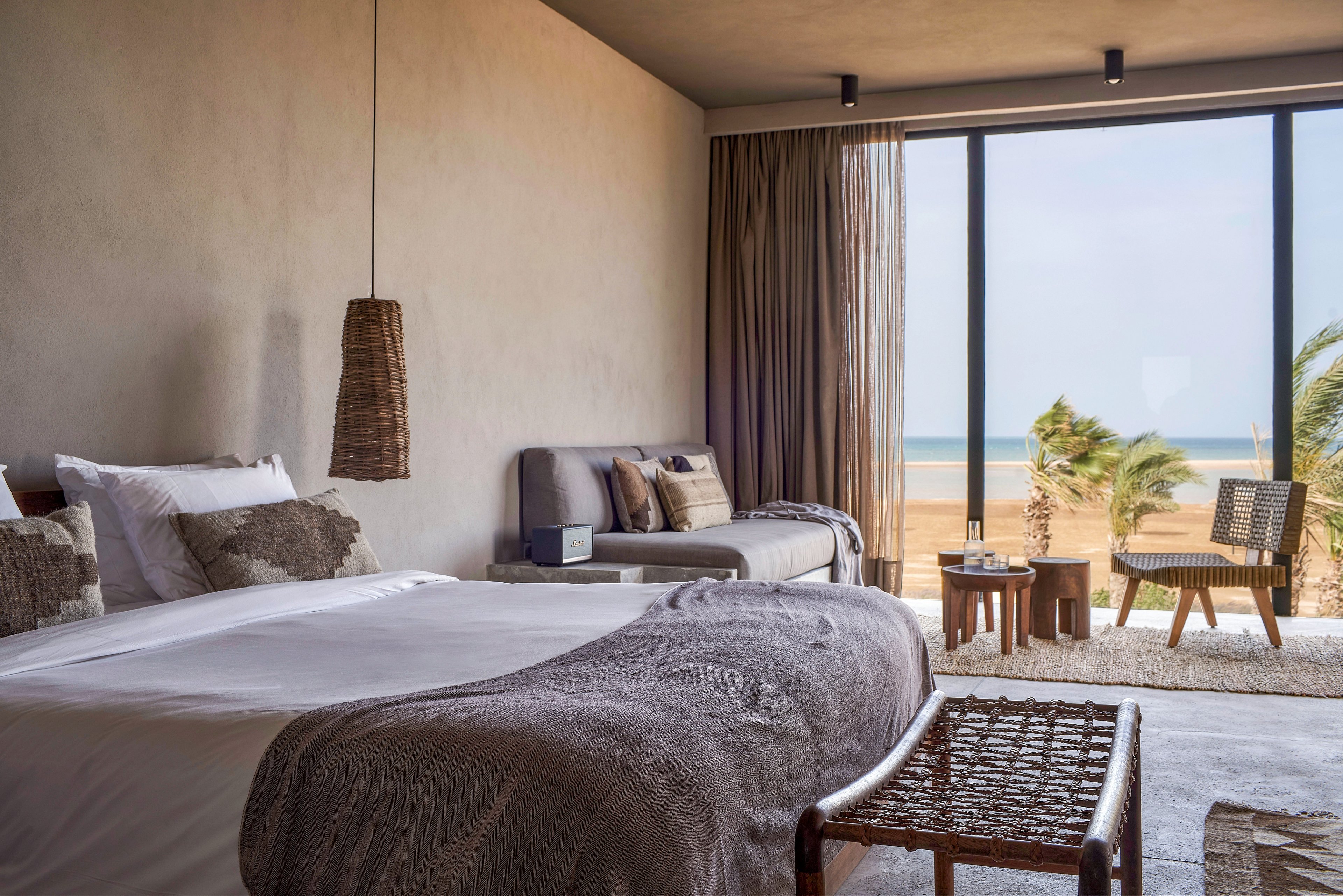 A stylish and cozy bedroom featuring a large bed with neutral-toned bedding, a woven pendant light, and a seating area. The room opens to a balcony with a stunning ocean view, palm trees, and outdoor furniture. The decor emphasizes natural materials and a minimalist aesthetic.