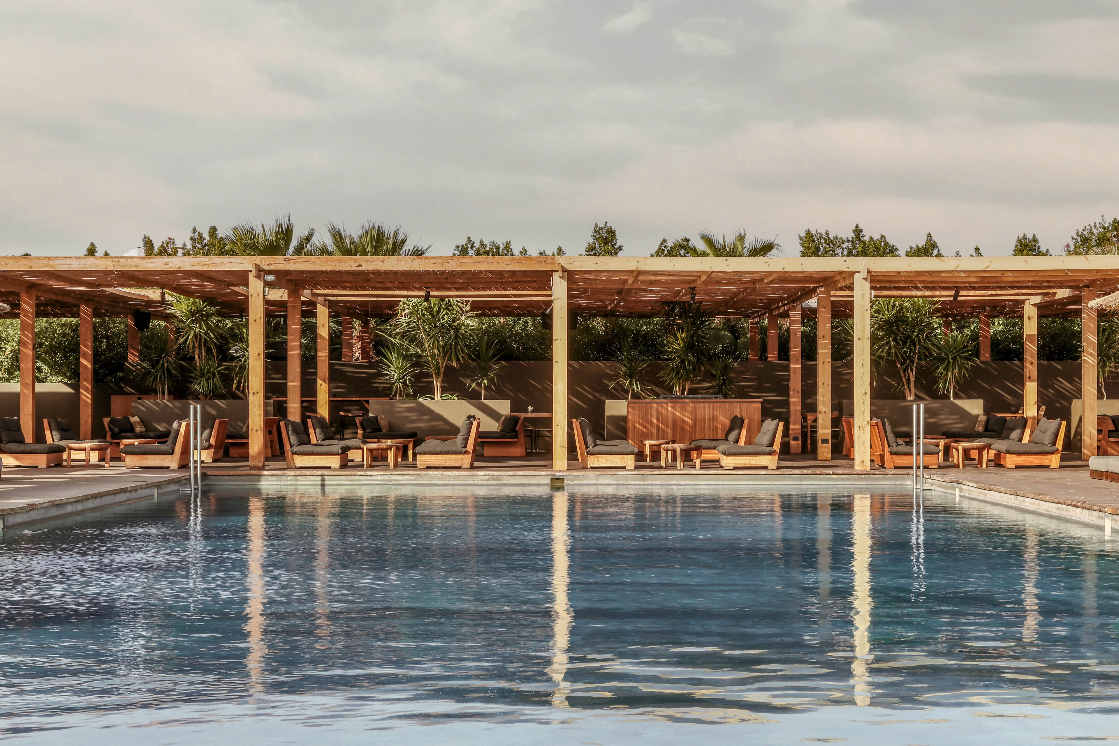 A serene outdoor pool surrounded by wooden cabanas and lush greenery under a partly cloudy sky. The setting exudes a luxurious and tranquil atmosphere, perfect for relaxation. The reflections on the water enhance the calm and inviting ambiance.