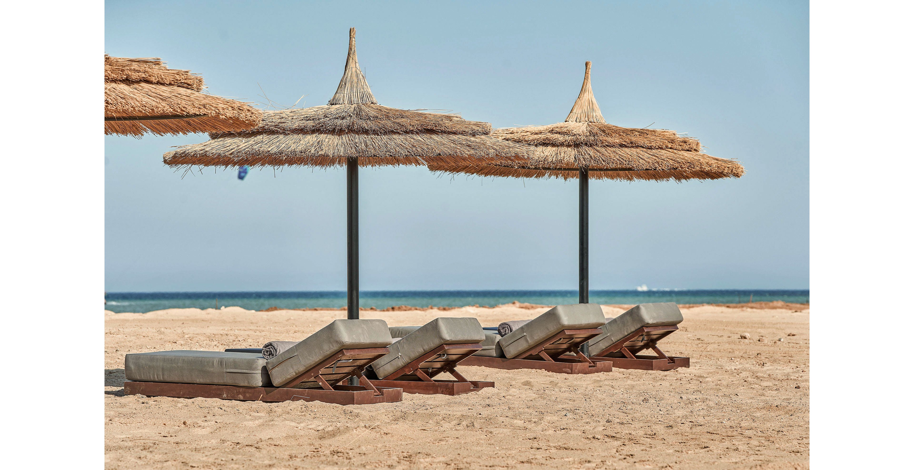 A serene beach setting featuring four cushioned loungers arranged under straw umbrellas. The sandy shore stretches out to meet the calm blue sea under a clear sky. The scene evokes relaxation and leisure in a tropical or coastal environment.