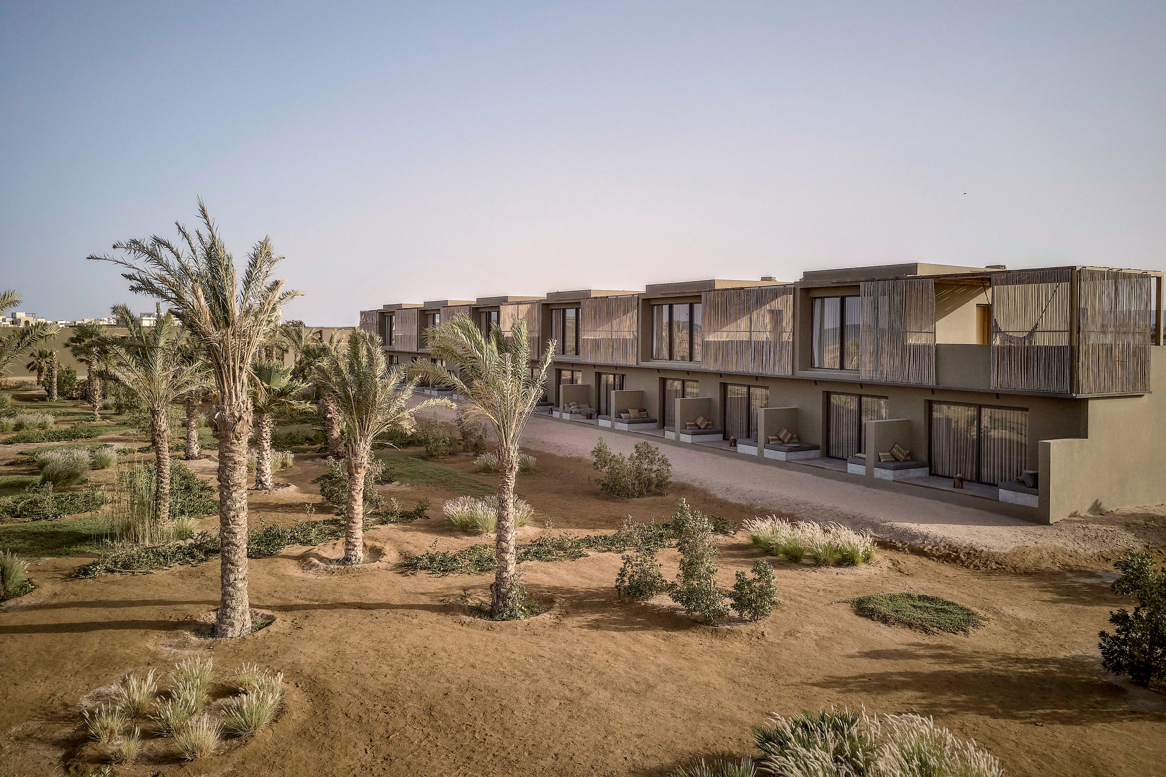 A contemporary resort featuring minimalist architecture set in a desert landscape. The building showcases clean lines and natural materials, surrounded by palm trees and sparse vegetation. The warm, earthy tones of the setting complement the serene and tranquil atmosphere.