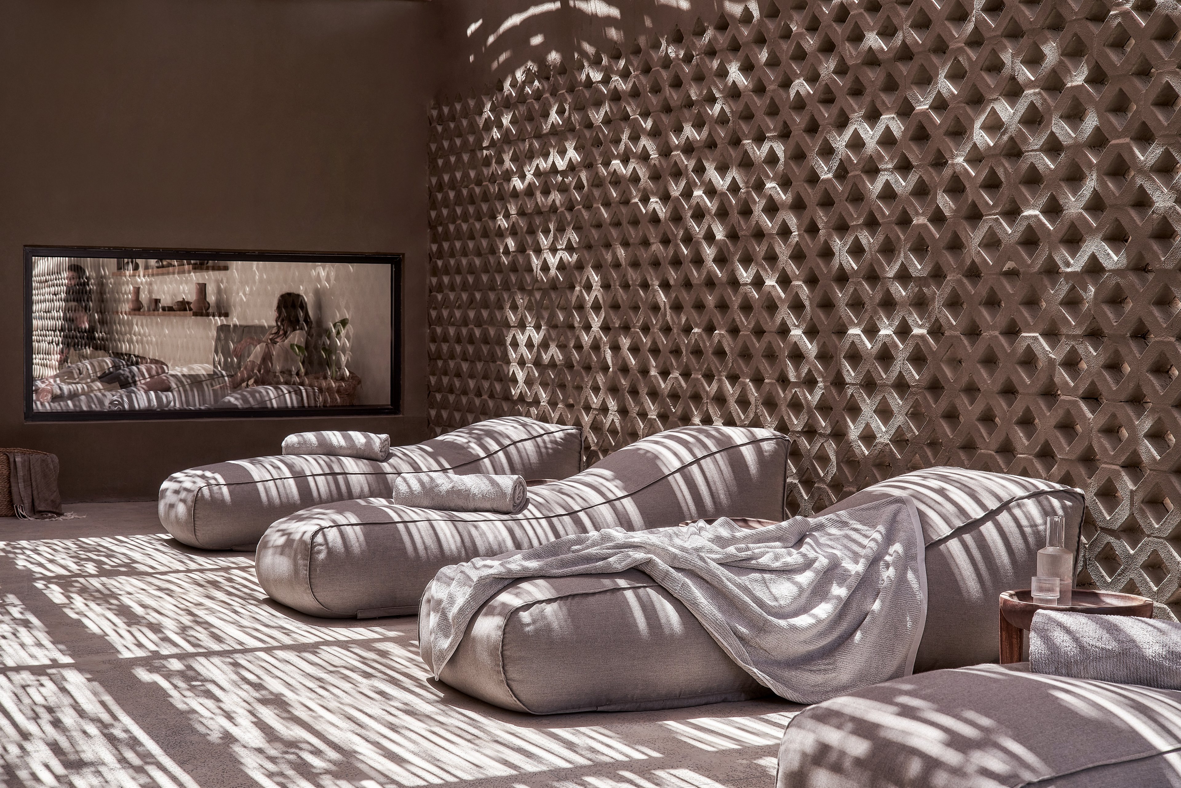 A serene lounge area featuring modern, cushioned recliners arranged in a tranquil setting. The space is illuminated by soft, patterned sunlight filtering through a decorative wall. A cozy blanket and a small side table with a candle enhance the relaxing ambiance.