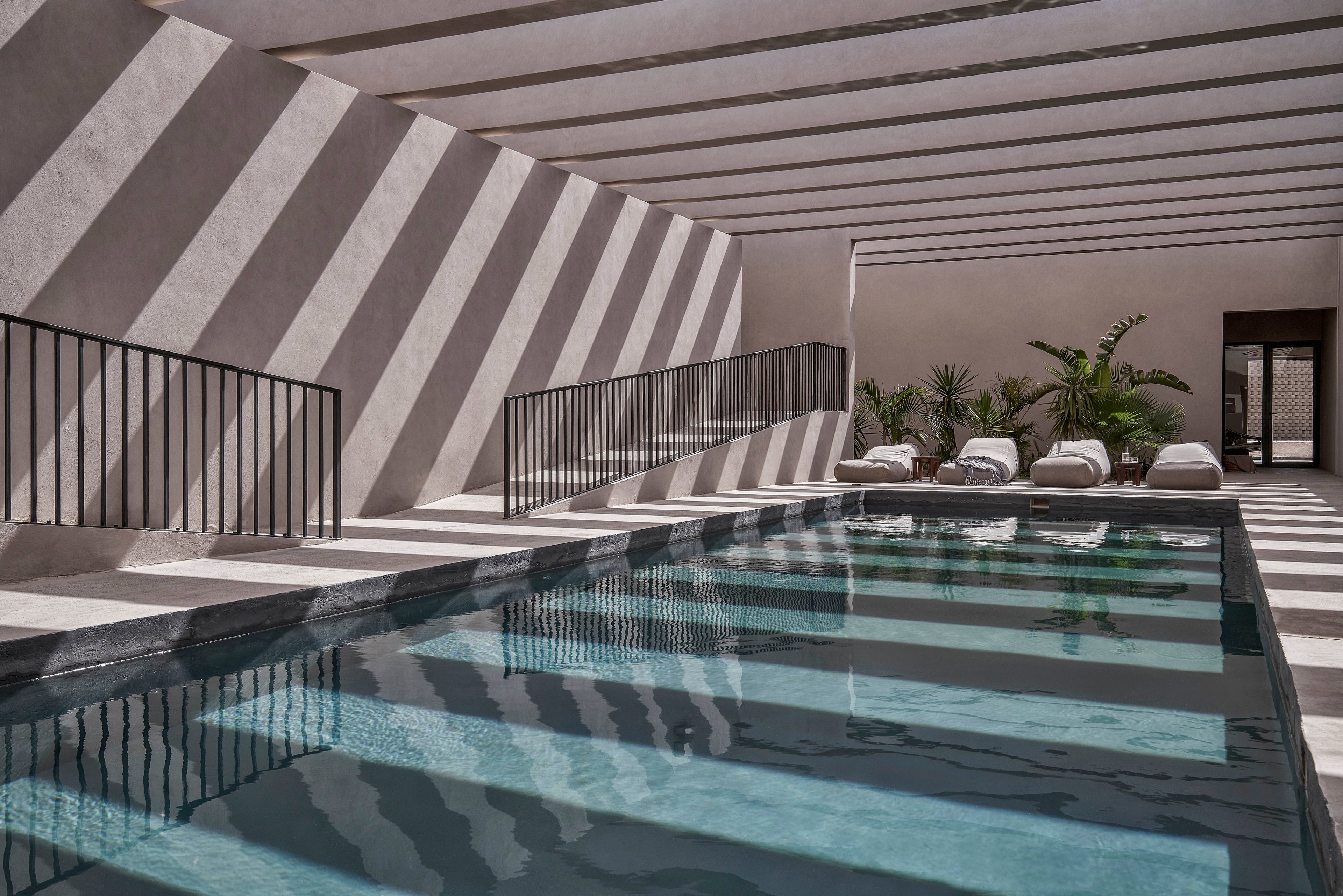 A sleek indoor pool surrounded by minimalist architecture, featuring striking striped shadows from overhead beams. The setting includes white lounge chairs and lush green plants, creating a serene and contemporary atmosphere. The design emphasizes clean lines and natural light.