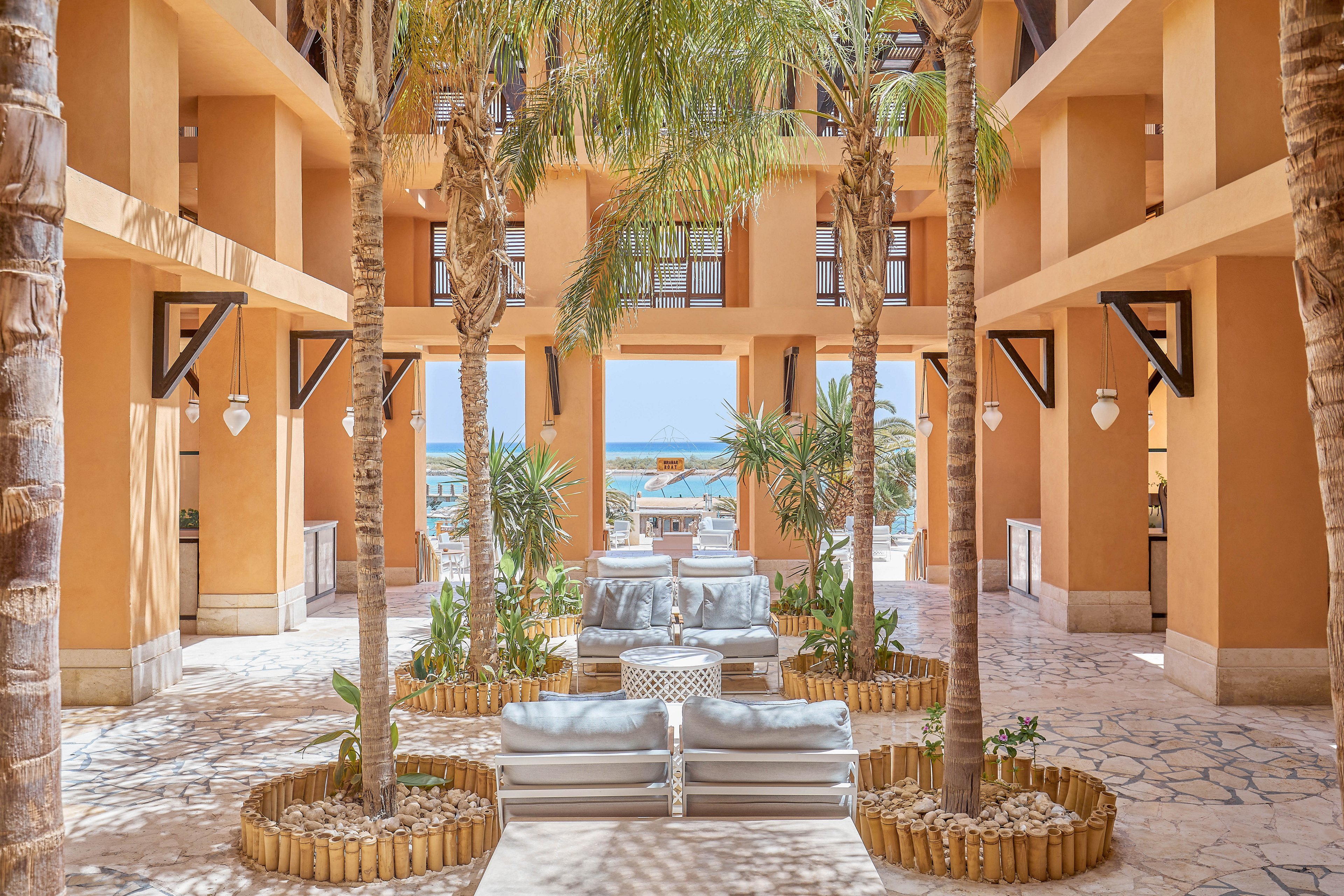 A serene courtyard in a luxury resort featuring tall palm trees, modern seating arrangements, and warm terracotta architecture. The open space leads to a view of the ocean, creating a tranquil and inviting atmosphere. Natural light enhances the earthy tones and tropical ambiance.