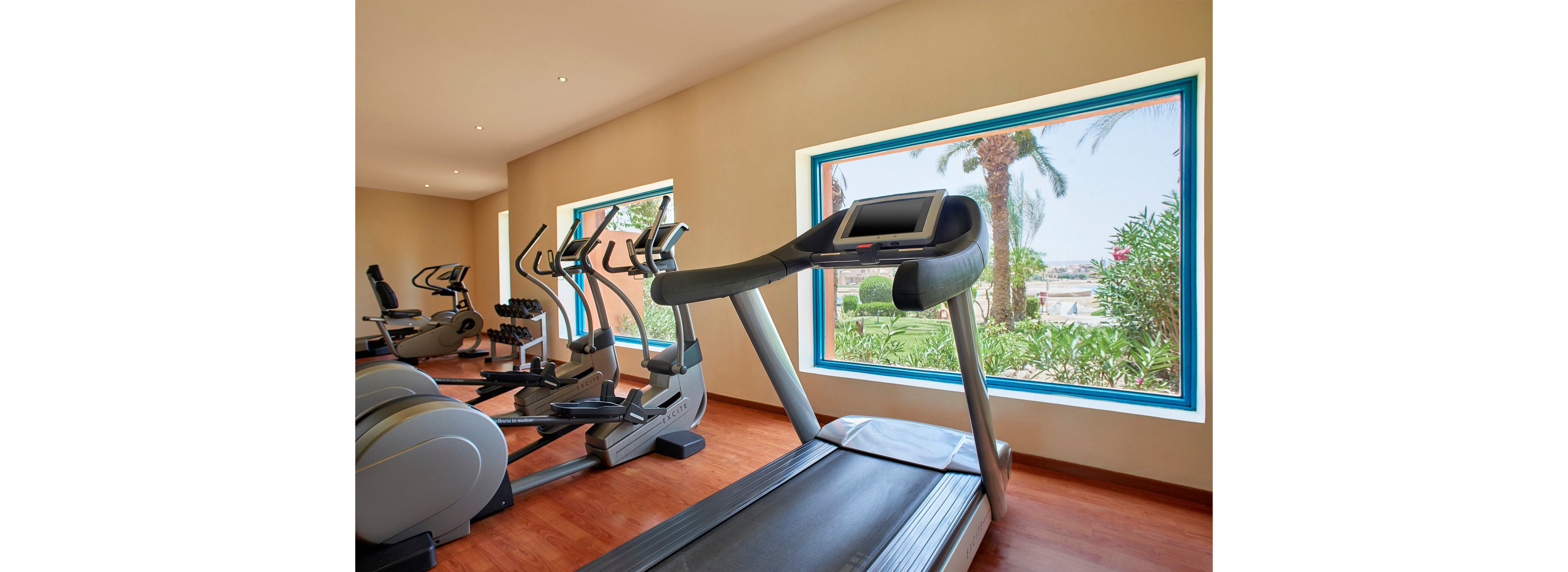 A bright and spacious gym featuring treadmills and exercise machines. Large windows provide natural light and a view of greenery outside. The wooden flooring adds warmth to the contemporary fitness setting.