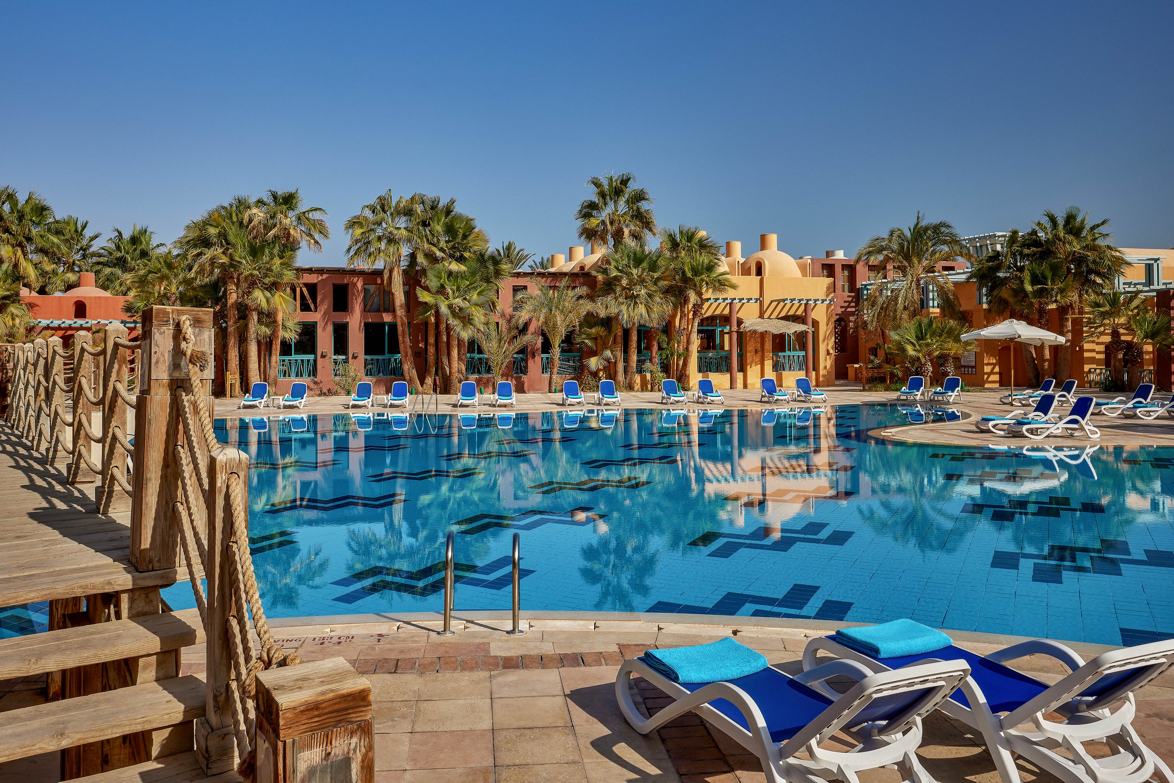A serene outdoor pool surrounded by palm trees and colorful resort buildings under a clear blue sky. The pool area features multiple lounge chairs with blue cushions and a wooden bridge crossing over the water. The setting exudes a tropical and relaxing vacation vibe.