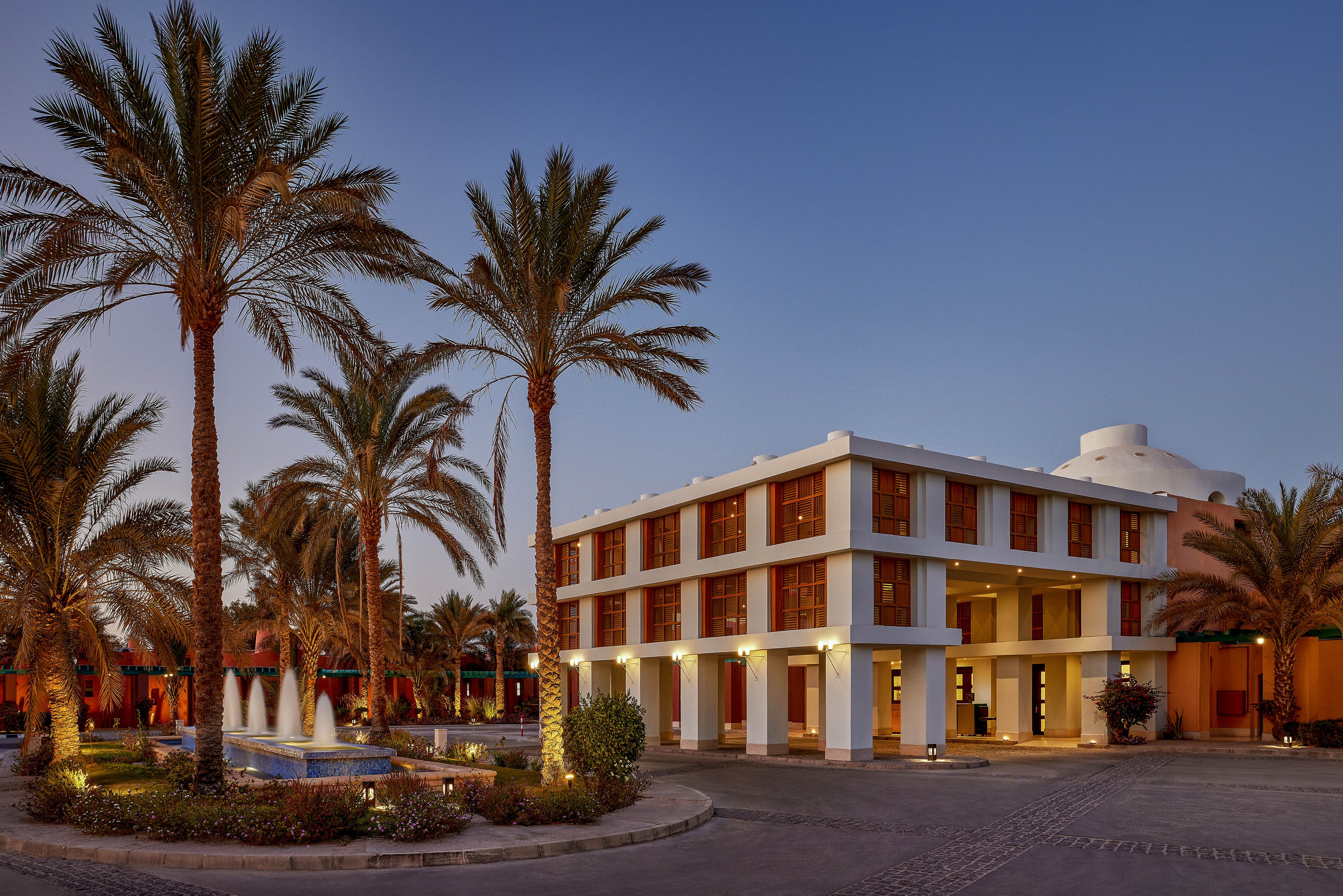 A contemporary hotel building surrounded by tall palm trees at dusk. The setting features a well-lit facade with large windows and a central fountain in the foreground. The warm lighting and clear sky create a serene and inviting atmosphere.