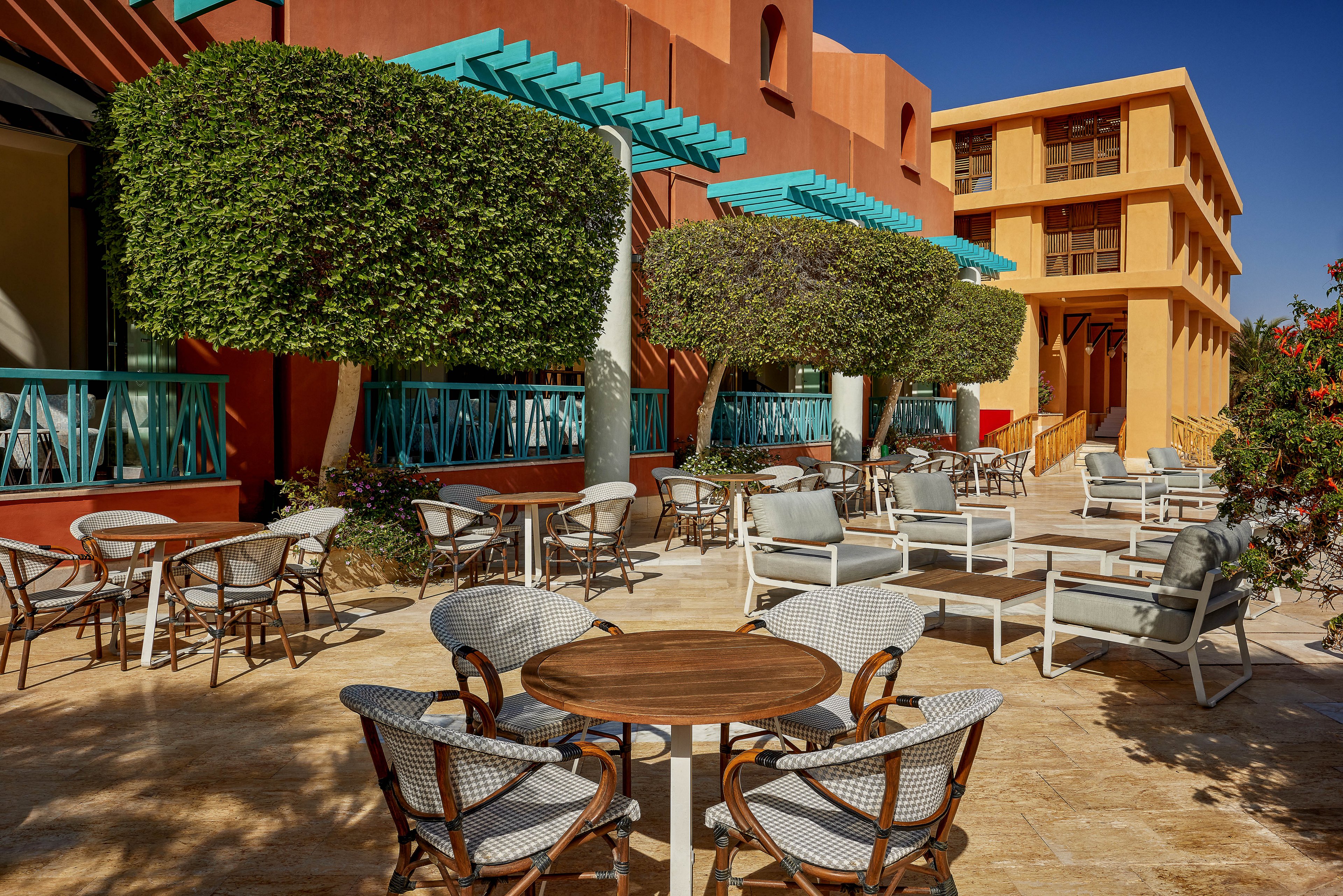 A spacious outdoor seating area featuring modern furniture and vibrant architecture. The setting includes neatly trimmed trees, colorful building facades, and shaded seating arrangements. The bright sunlight enhances the warm tones of the space, creating a welcoming atmosphere.