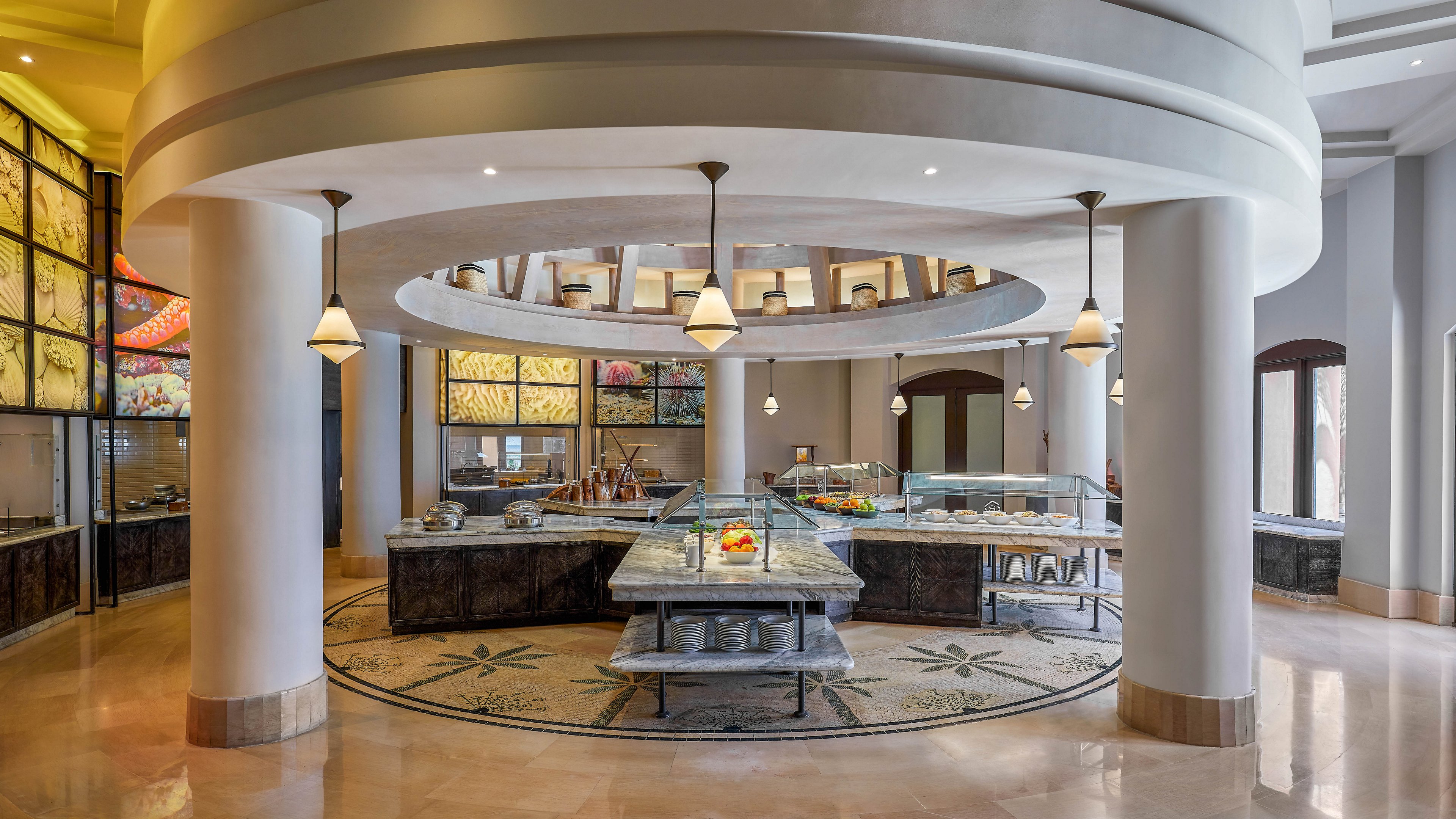 A spacious and well-lit buffet dining area featuring a circular layout with modern architectural elements. The central buffet station is surrounded by decorative columns and illuminated by hanging pendant lights. The setting includes a variety of food displays and a polished floor with intricate patterns, creating a sophisticated ambiance.