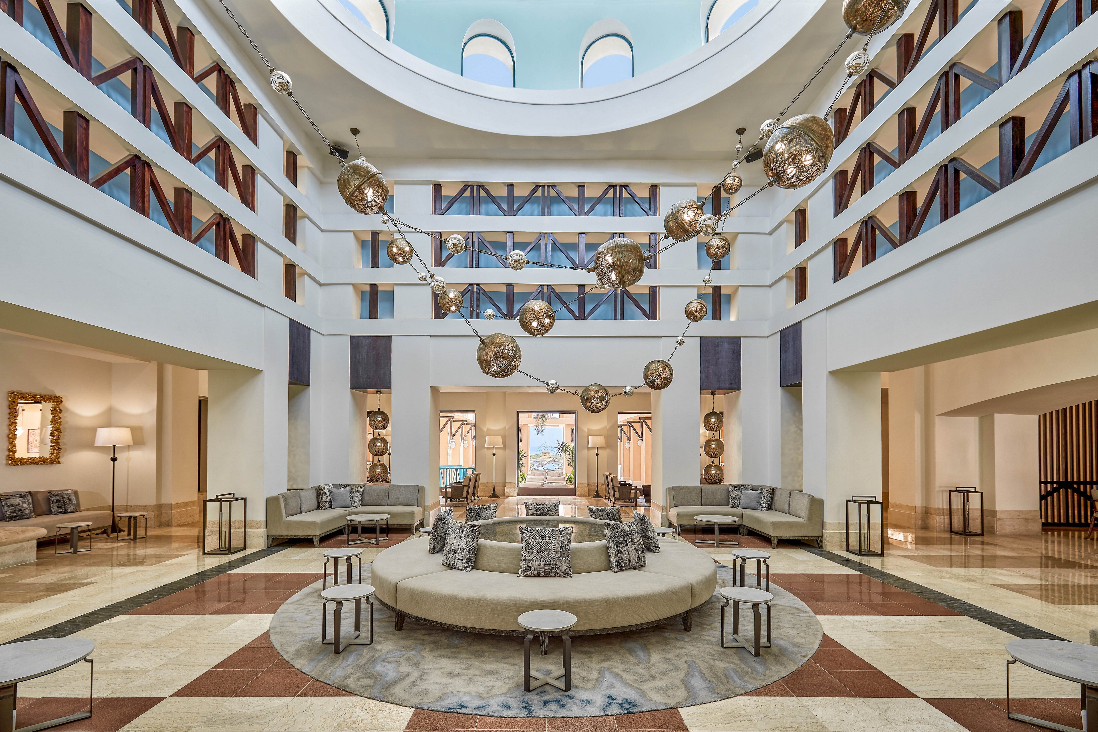 A spacious hotel lobby featuring a circular seating arrangement surrounded by modern furniture. The ceiling showcases a skylight with arched windows, complemented by hanging spherical light fixtures. Neutral tones and geometric patterns create a sophisticated ambiance.