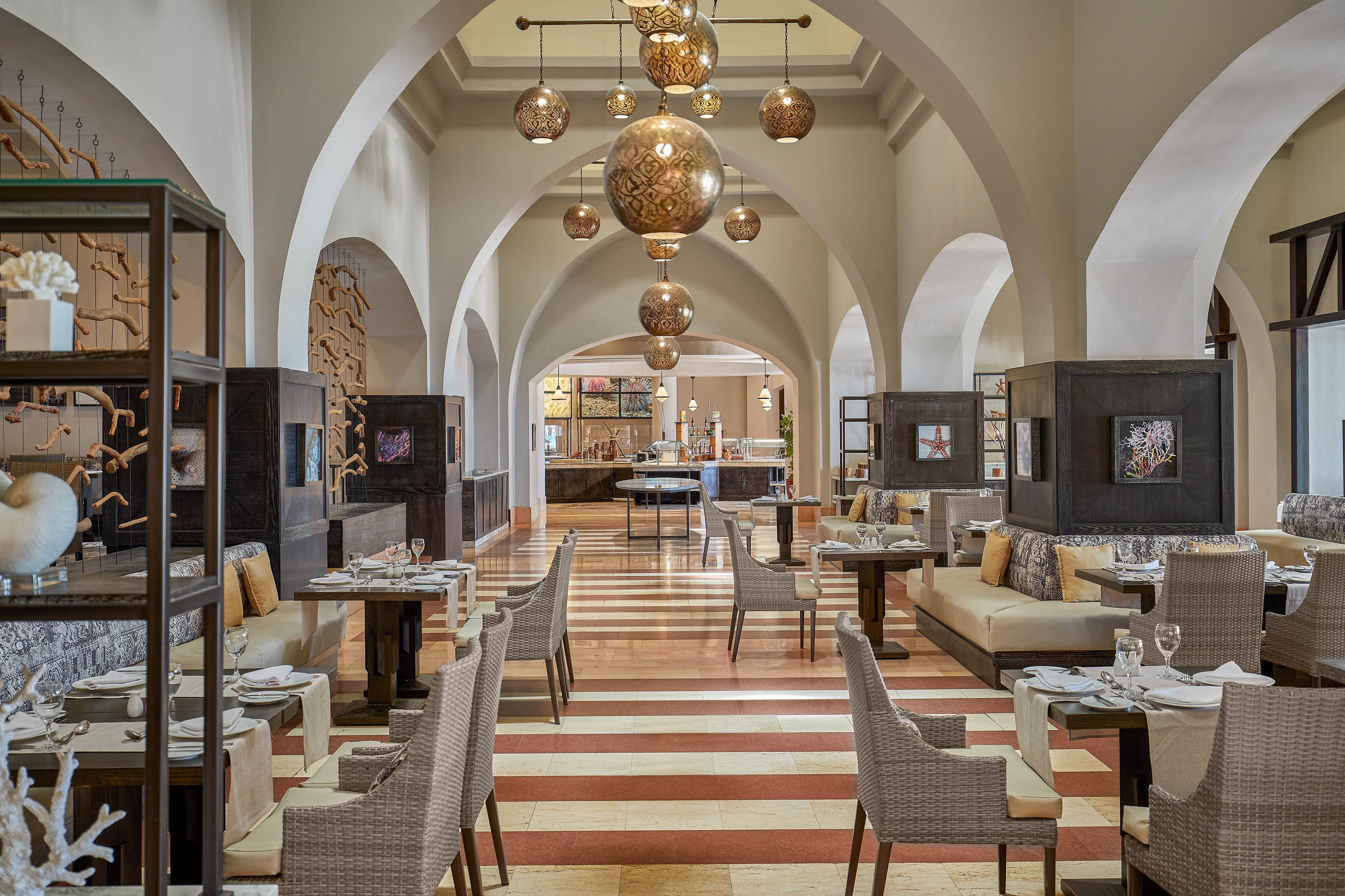 A spacious restaurant with Mediterranean-inspired decor featuring arched ceilings, hanging lanterns, and neutral tones. The seating area includes woven chairs and cushioned benches arranged around wooden tables. The warm lighting and intricate details create a sophisticated and inviting ambiance.