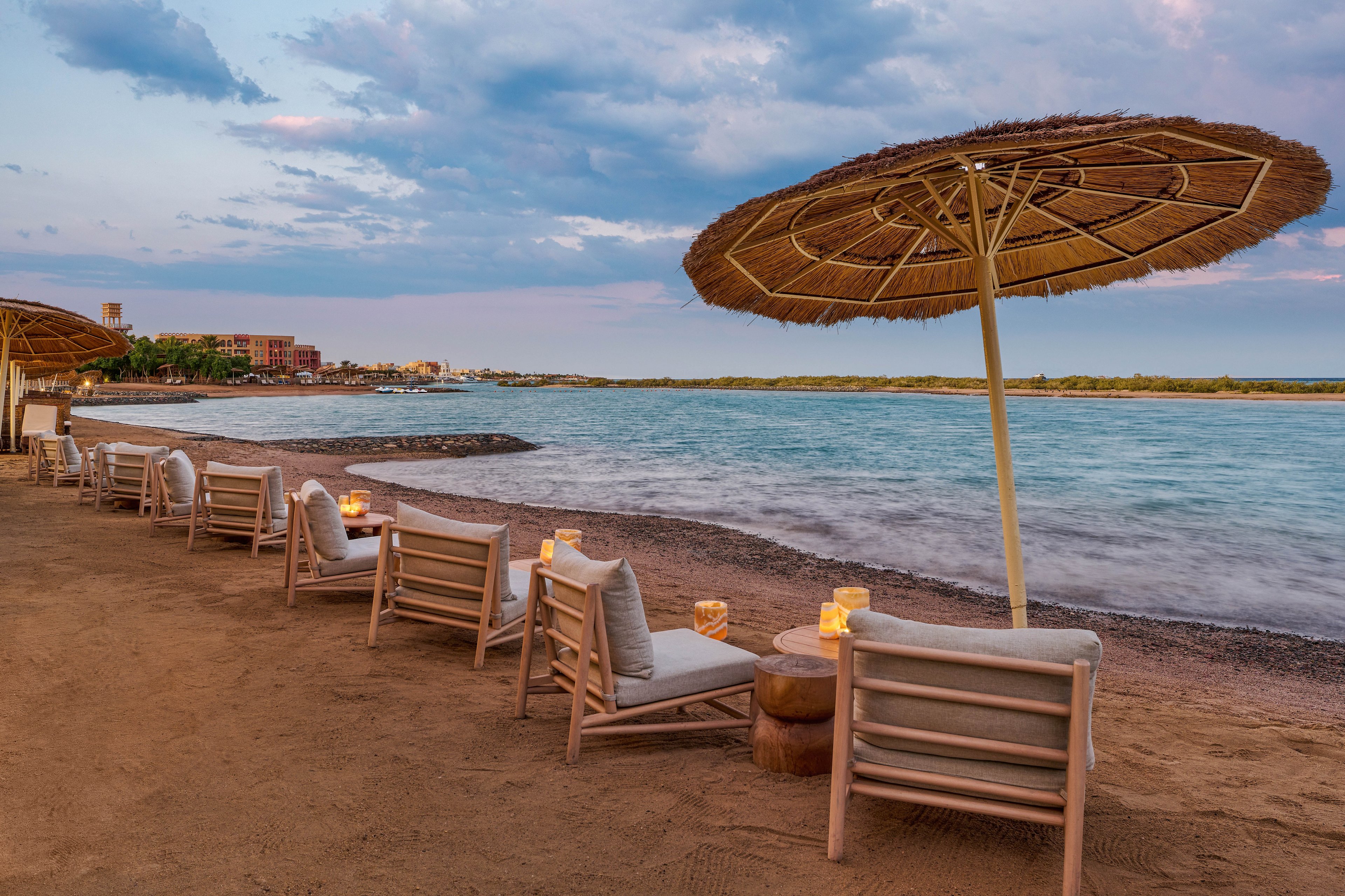A serene beachfront setting featuring wooden chairs with cushions arranged under straw umbrellas. The view includes calm waters and a distant shoreline, with soft lighting from candles on small tables. The sky is painted with pastel hues, creating a tranquil and inviting atmosphere.