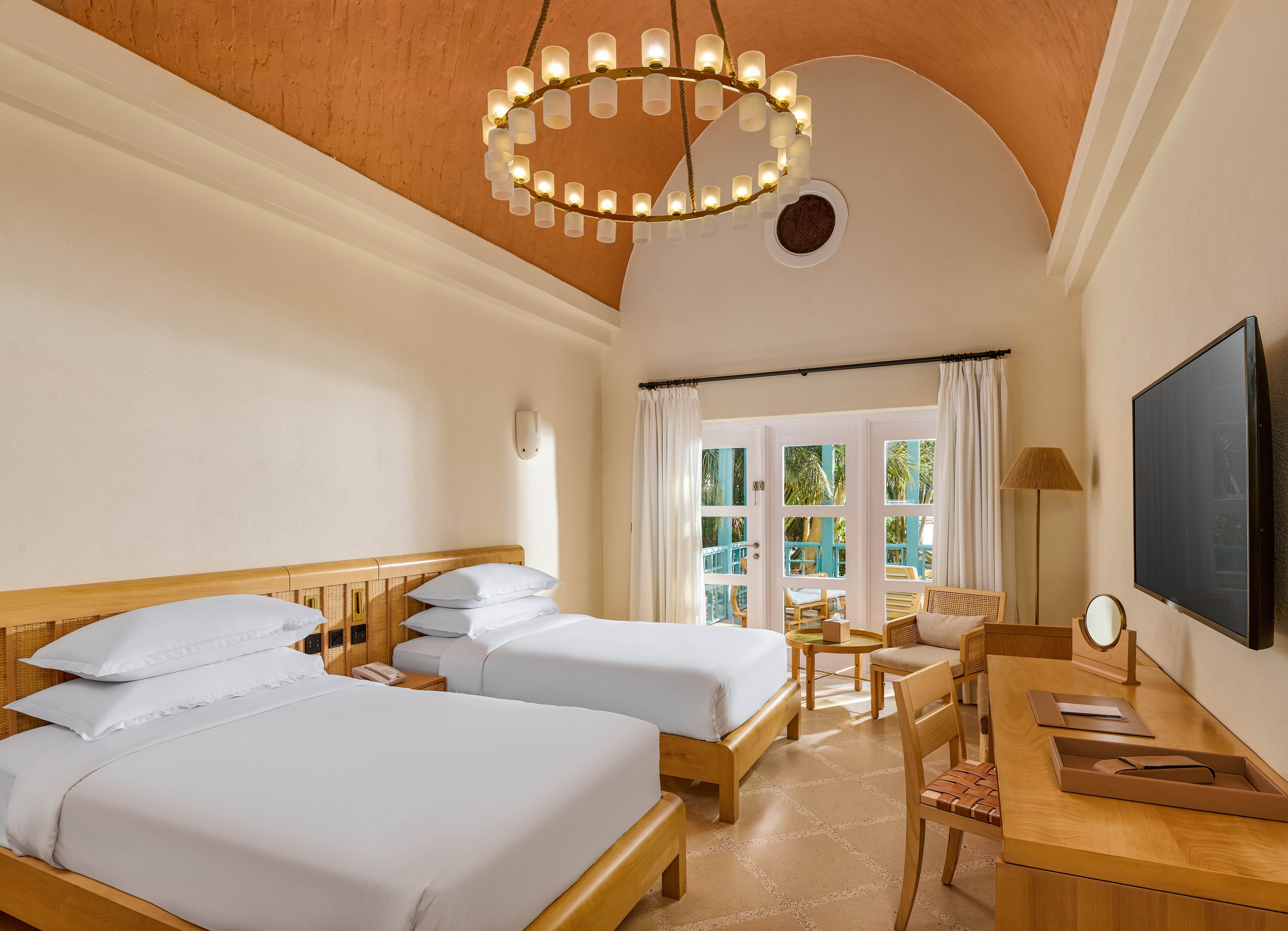 A spacious hotel room featuring two neatly made twin beds with white linens. The room is adorned with warm wooden furniture, a large chandelier, and a vaulted ceiling. A flat-screen TV is mounted on the wall, and a seating area is positioned near large windows that overlook a lush outdoor view. The decor is minimalistic and inviting.