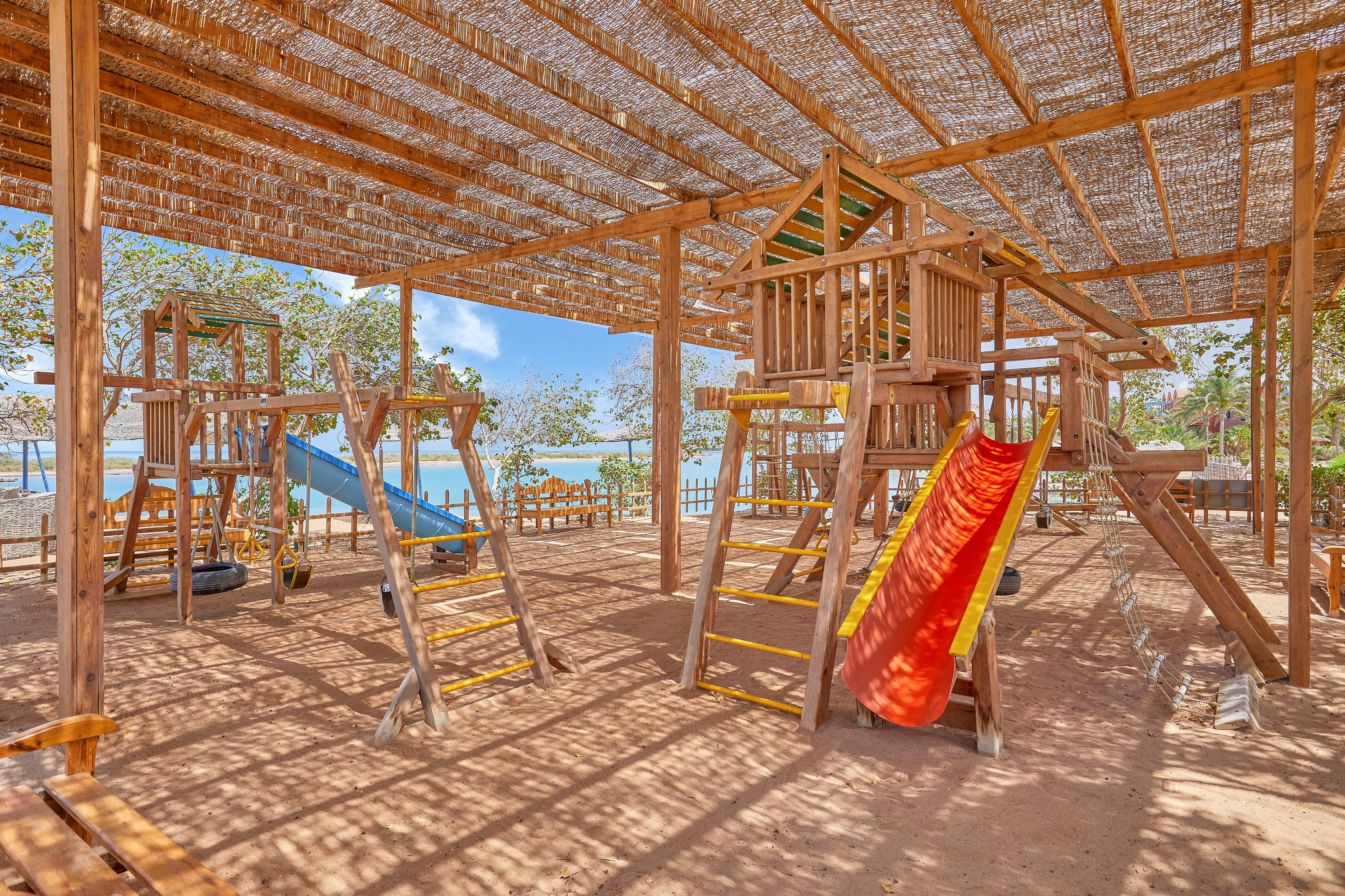 A spacious outdoor playground featuring wooden climbing structures, slides, and swings under a shaded canopy. The setting is bright and sunny, with a view of trees and a distant body of water. The red and yellow slide adds a vibrant touch to the natural wooden tones.