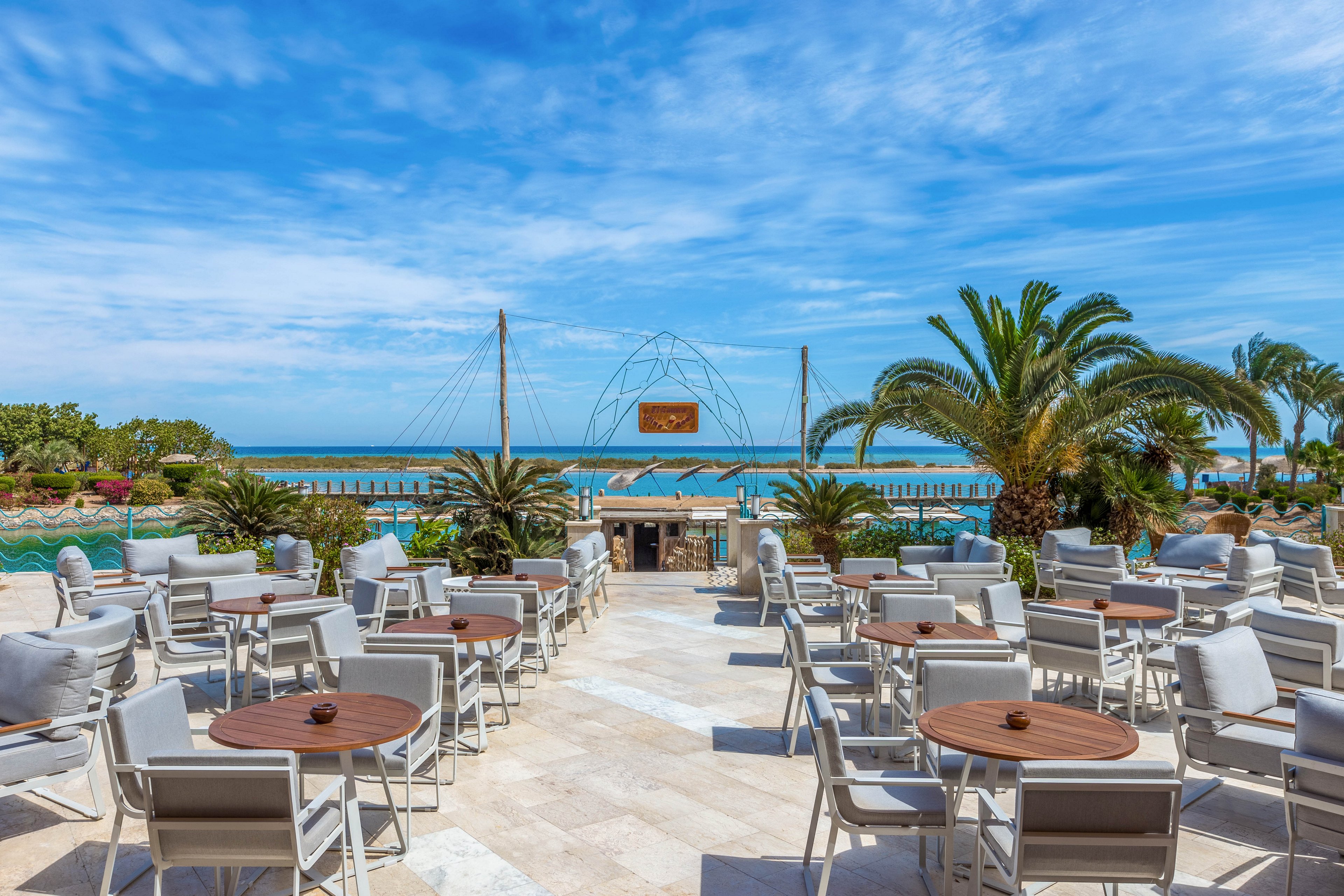 An open-air seating area featuring numerous tables and chairs arranged on a tiled patio. The setting overlooks a vibrant blue ocean, framed by lush palm trees and greenery. The sky is clear with scattered clouds, creating a serene and inviting atmosphere.