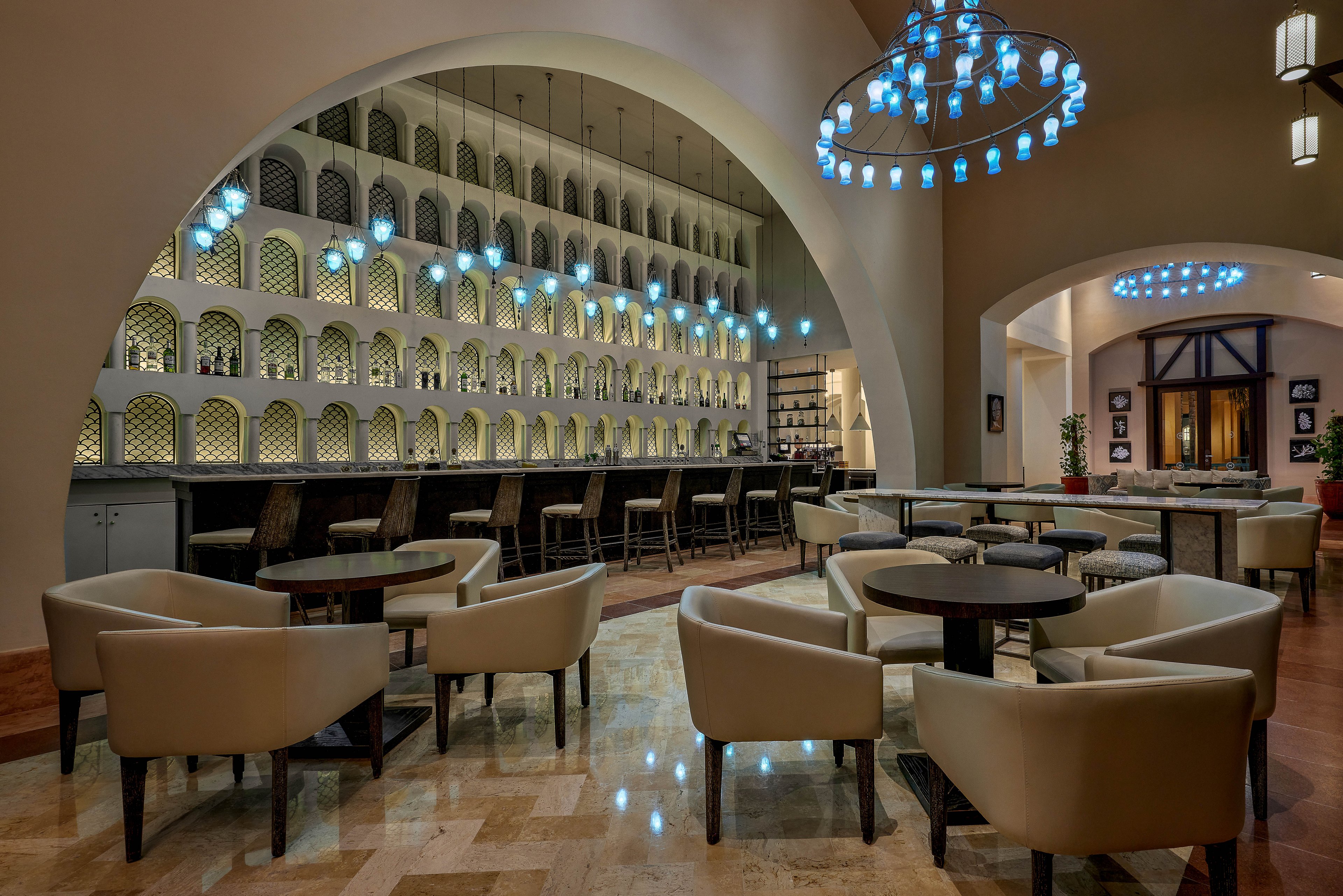 A stylish bar lounge featuring contemporary furniture and a unique wall design with illuminated shelving. The setting includes round tables, cushioned chairs, and a polished marble floor. Blue pendant lights and chandeliers add a sophisticated ambiance to the space.