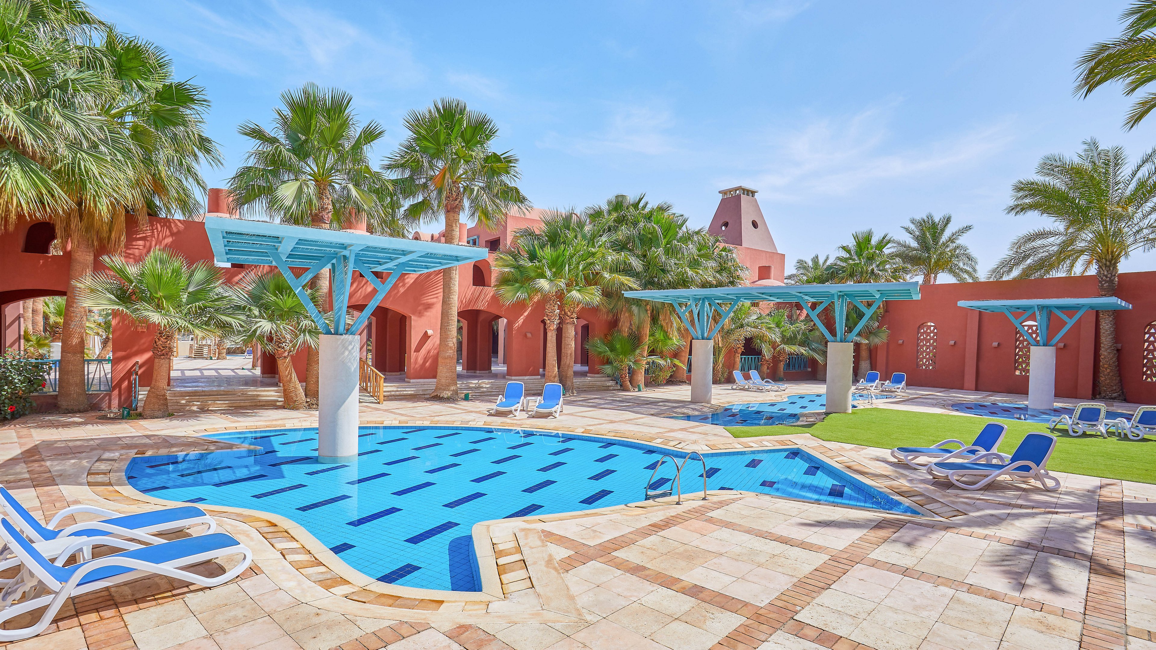 A vibrant outdoor pool surrounded by palm trees and red stucco buildings in a resort setting. Blue and white loungers are arranged around the pool, which features geometric tile patterns and shaded structures. The sunny atmosphere and lush greenery create a relaxing vacation vibe.