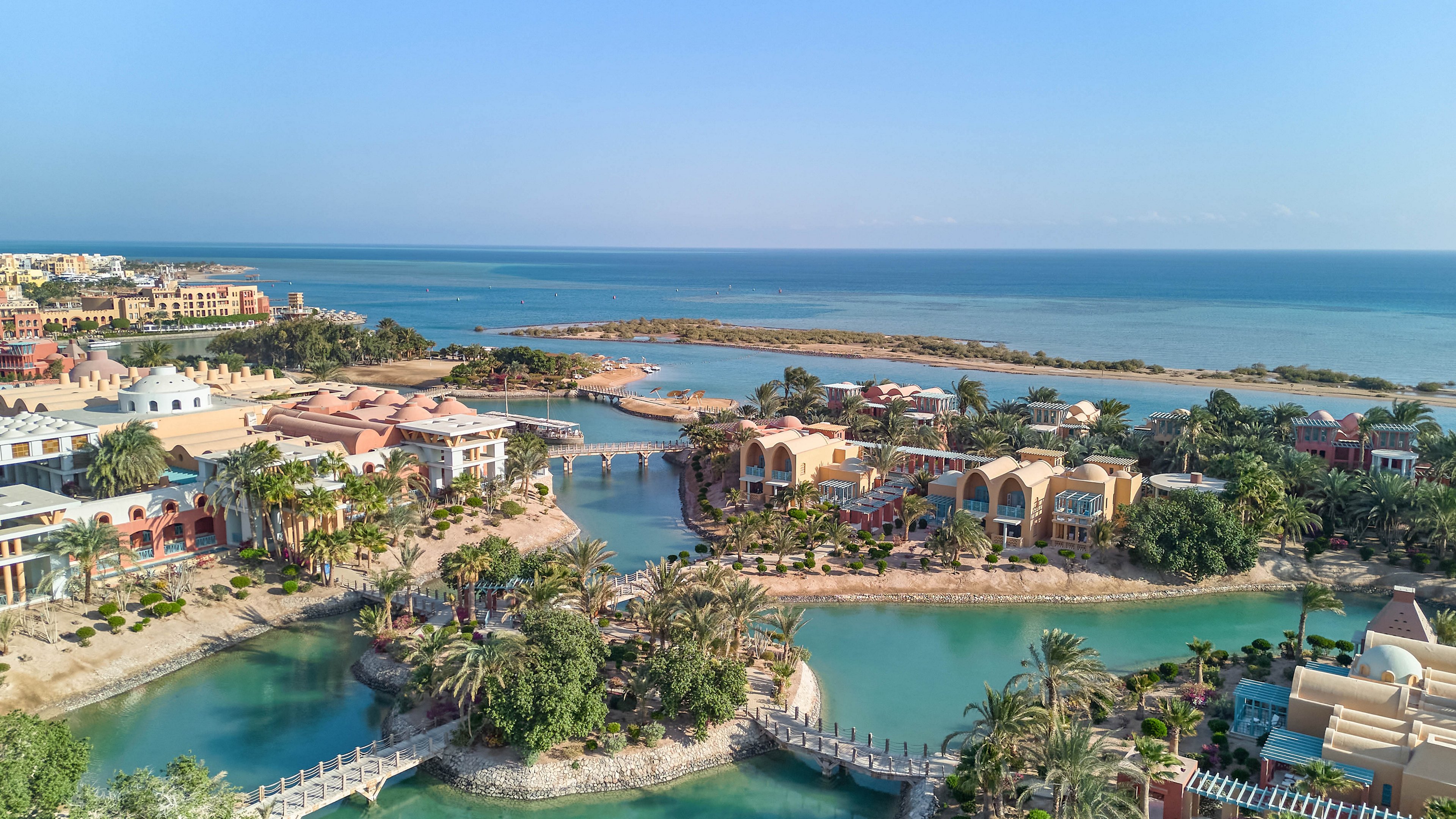 A picturesque coastal resort featuring vibrant architecture surrounded by lush greenery and tranquil lagoons. The setting overlooks a serene ocean, with bridges connecting various areas of the resort. The bright, sunny atmosphere highlights the tropical environment and inviting waters.