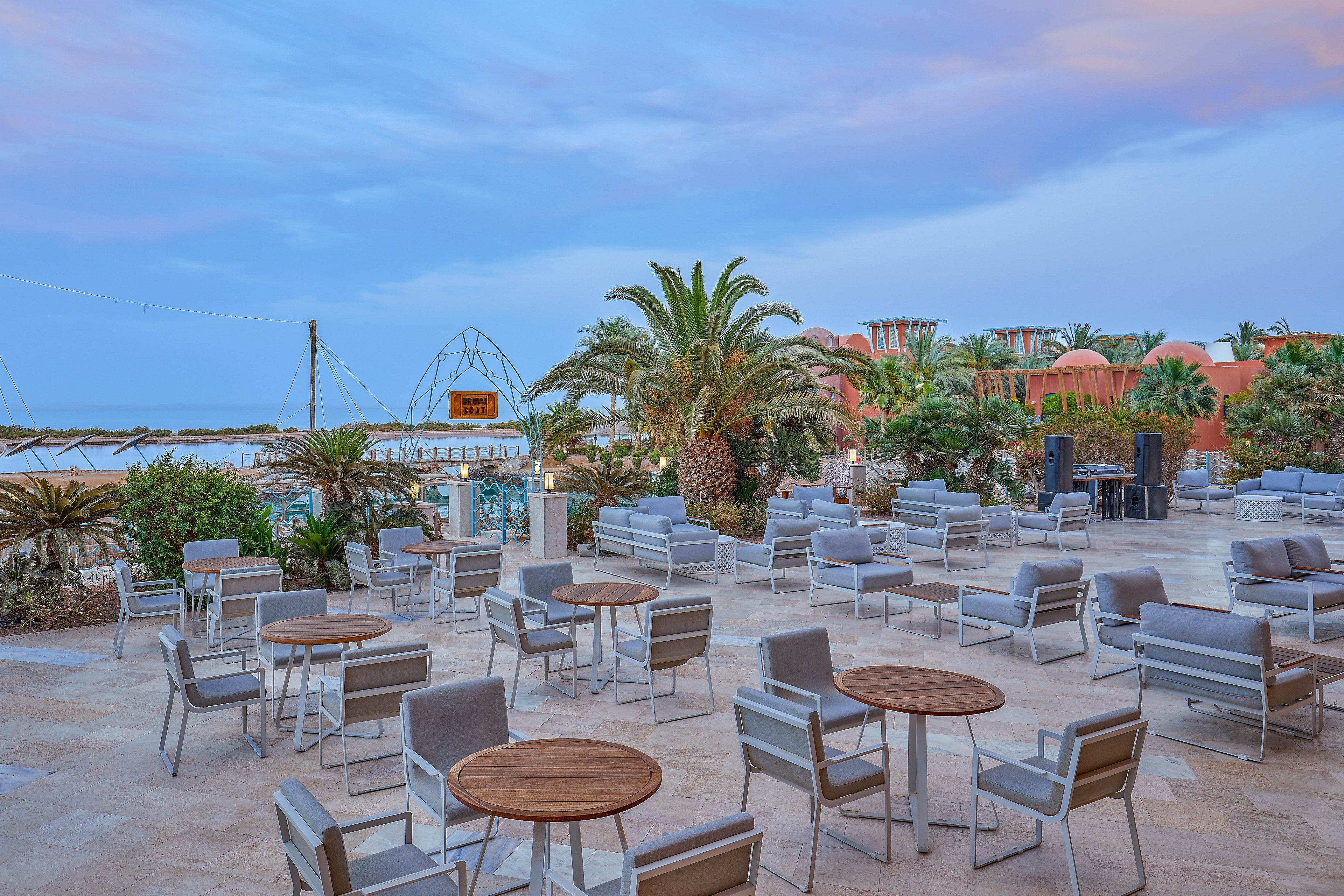 An open-air seating area featuring multiple tables and chairs arranged on a tiled patio. Surrounded by lush palm trees and greenery, the setting overlooks a serene waterfront under a soft evening sky. The atmosphere is tranquil and inviting, ideal for relaxation or casual gatherings.