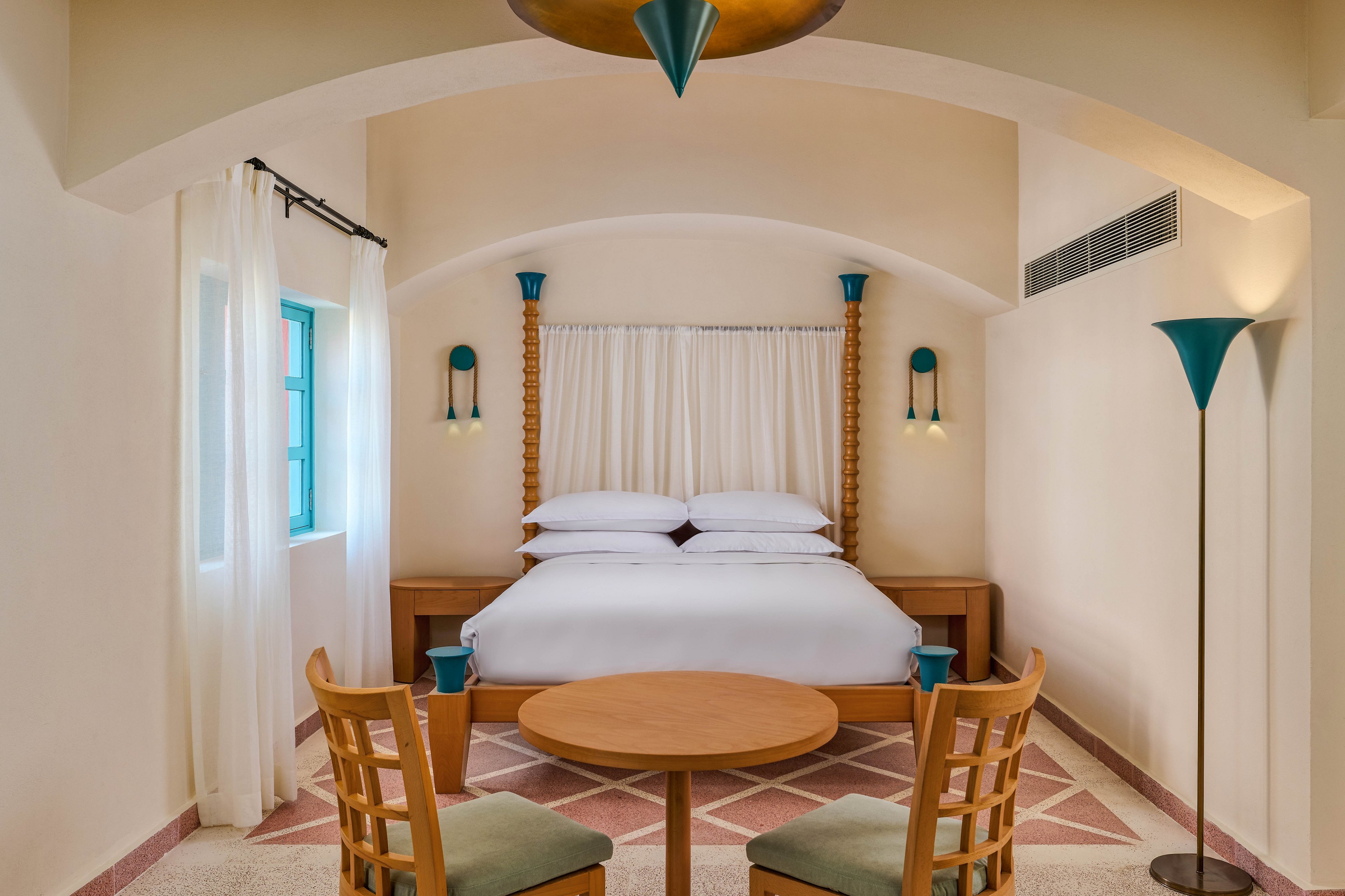 A well-lit hotel room featuring a comfortable double bed with white linens and a canopy-style headboard. The room includes a small round wooden table with two chairs, teal accents, and soft lighting. The arched ceiling and pastel tones create a warm and inviting atmosphere.