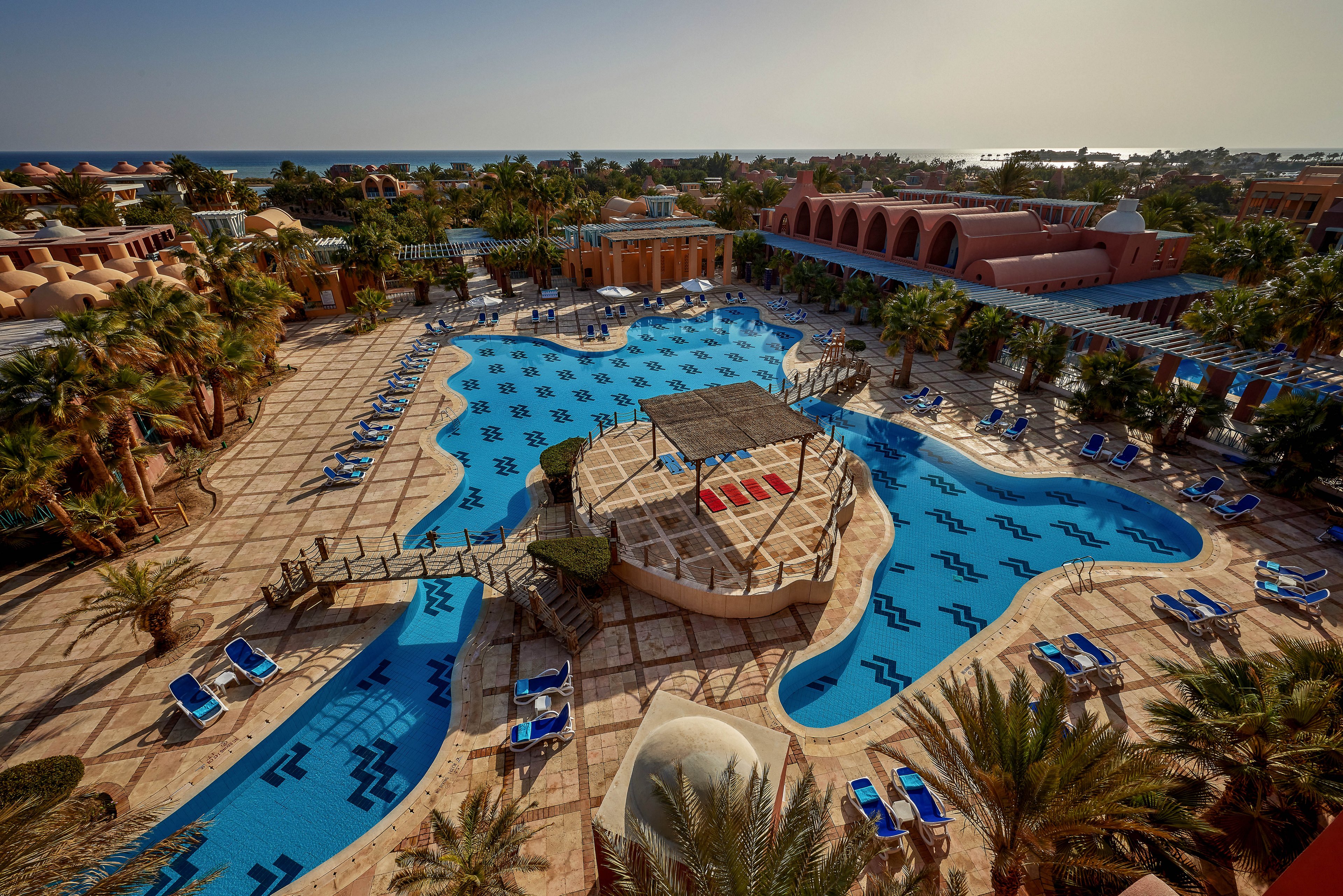 A large resort pool surrounded by lounge chairs and palm trees, set against a backdrop of the ocean. The pool features unique wave-like patterns and a central shaded seating area. The setting is sunny and tranquil, ideal for relaxation and leisure.