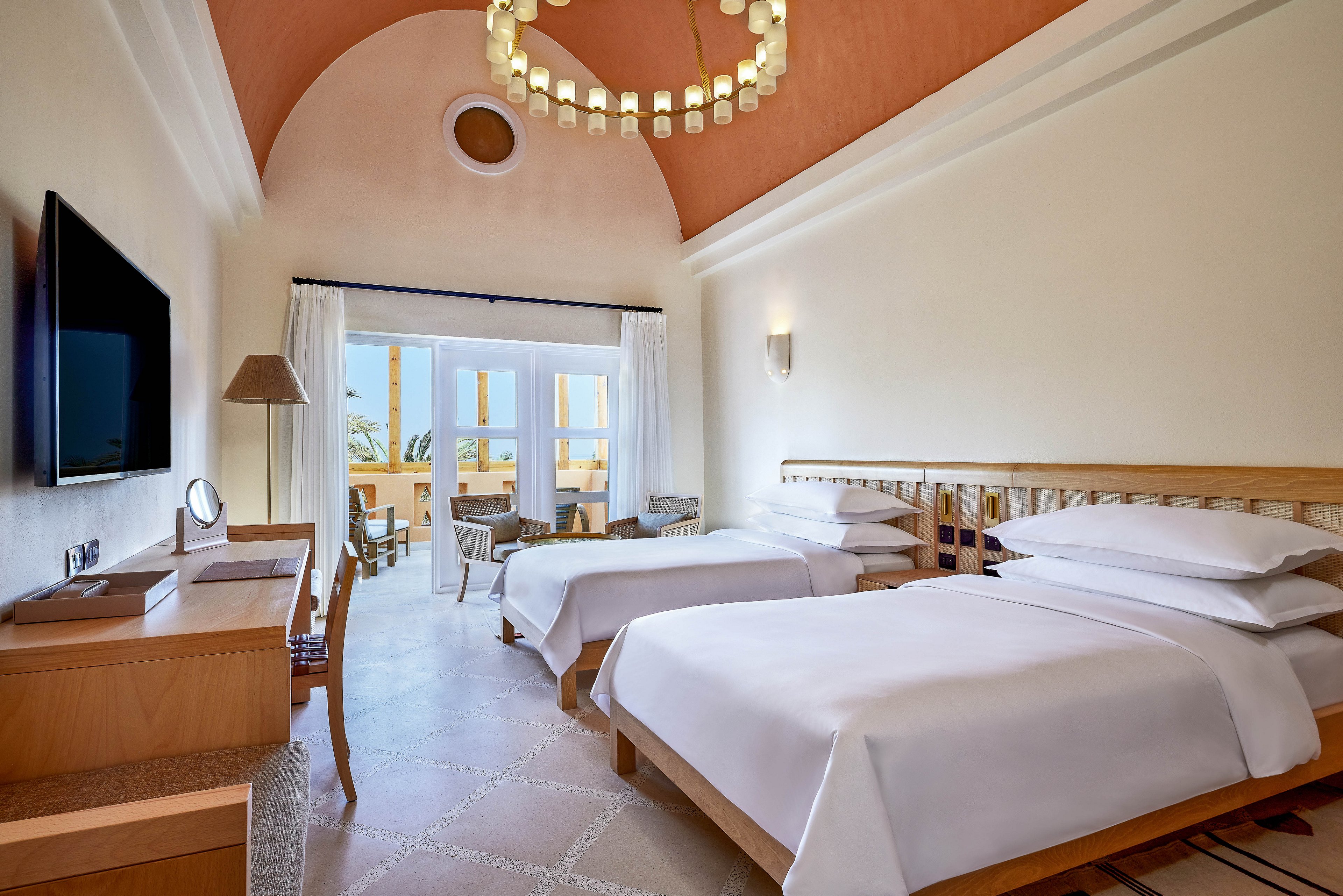 A spacious hotel room featuring two neatly arranged twin beds with white linens. The room includes wooden furniture, a wall-mounted TV, and a desk with a lamp. Large windows open to a balcony with outdoor seating, offering a view of palm trees and clear skies. The ceiling has a warm terracotta tone and a decorative chandelier.
