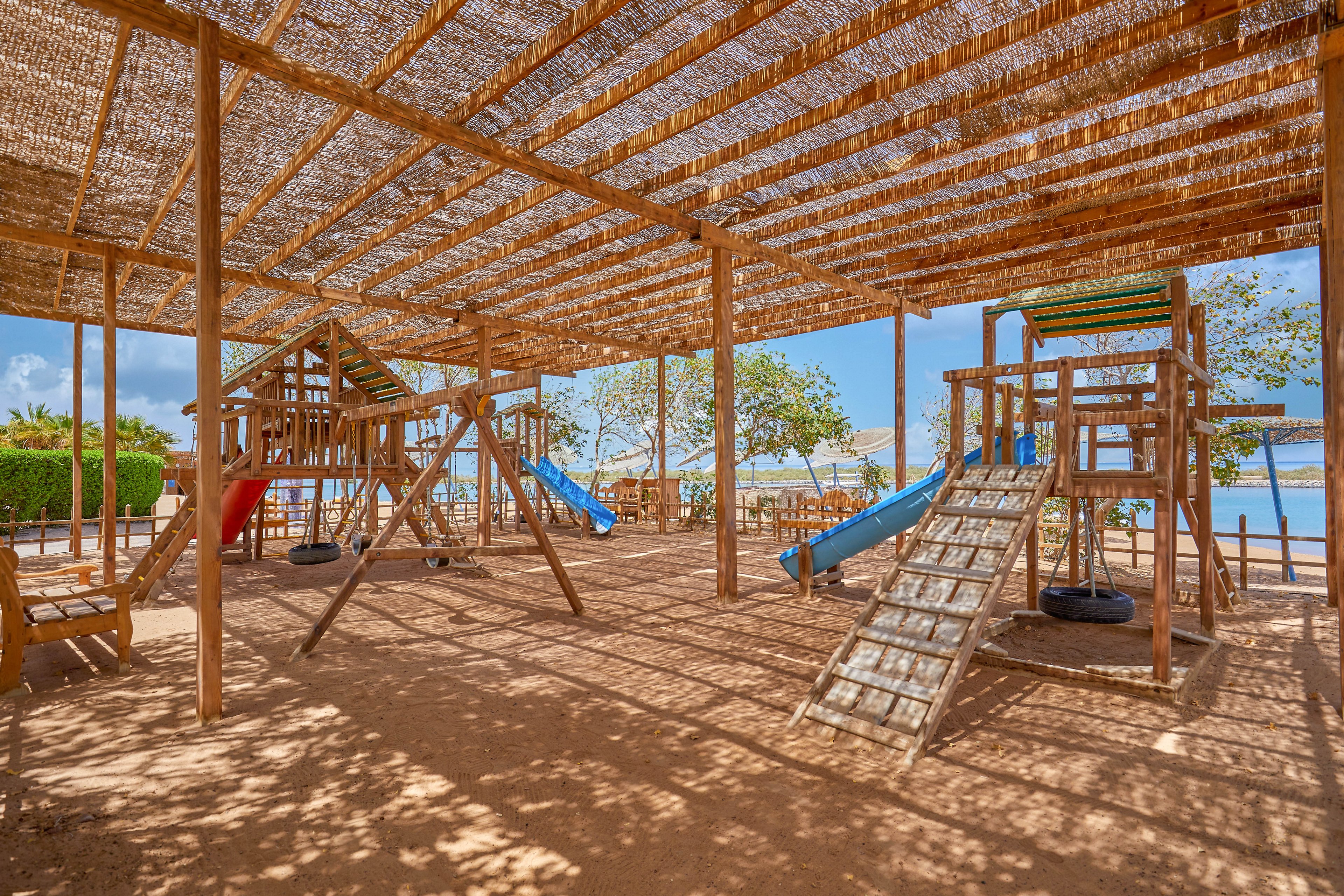 A spacious outdoor playground featuring wooden climbing structures, slides, and swings under a shaded canopy. The setting is bright and sunny, with trees and a waterfront visible in the background. The area is designed for children and offers a natural, rustic aesthetic.