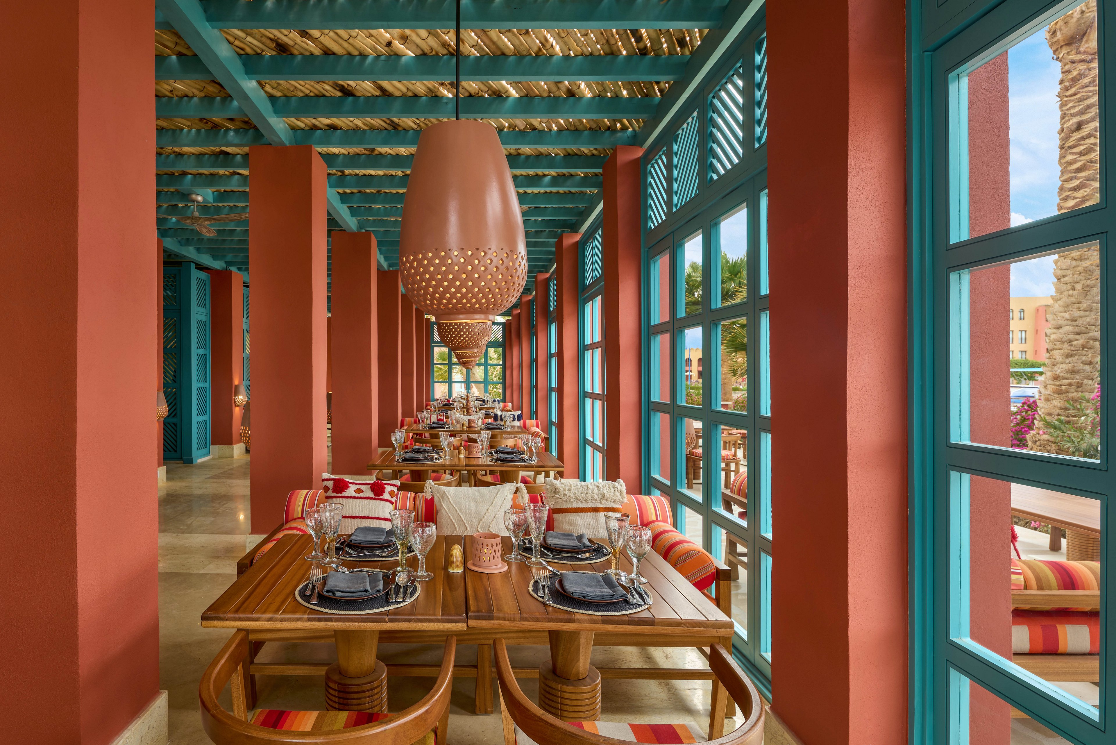A lively restaurant setting featuring wooden tables and chairs adorned with striped cushions. The space is framed by bold red walls, turquoise window frames, and hanging pendant lights. Natural light streams through large windows, creating a warm and inviting atmosphere.