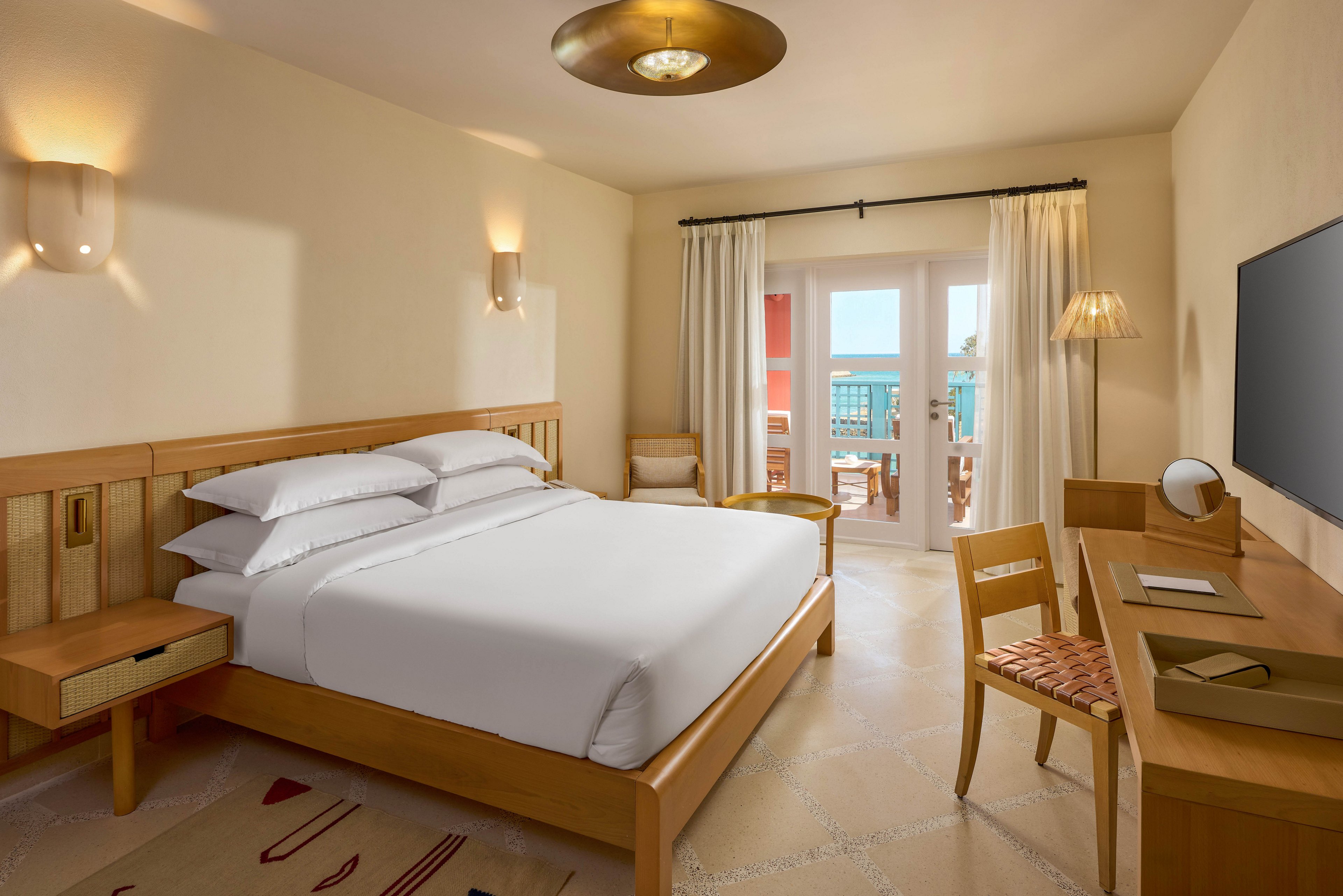 A bright and modern hotel room featuring a neatly made bed, wooden furniture, and a desk with a flat-screen TV. The room opens to a balcony with a view of colorful buildings and the outdoors. Warm lighting and neutral tones create a cozy and inviting atmosphere.