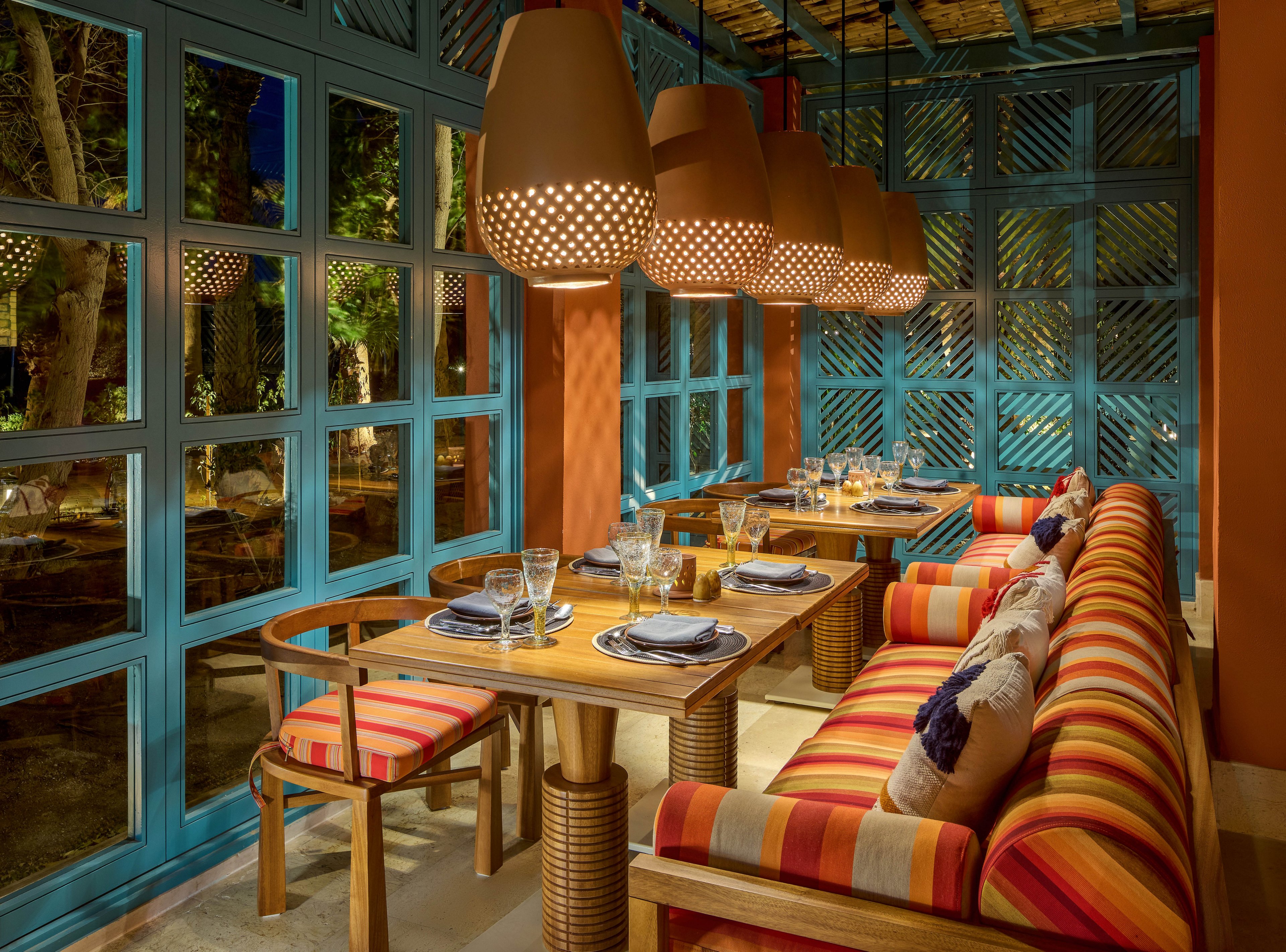 A warmly lit restaurant interior featuring colorful striped seating and wooden tables set with glassware and plates. The space is adorned with large, perforated pendant lights and blue-framed windows overlooking a lush outdoor area. The vibrant decor creates a cozy and inviting atmosphere, perfect for dining and social gatherings.