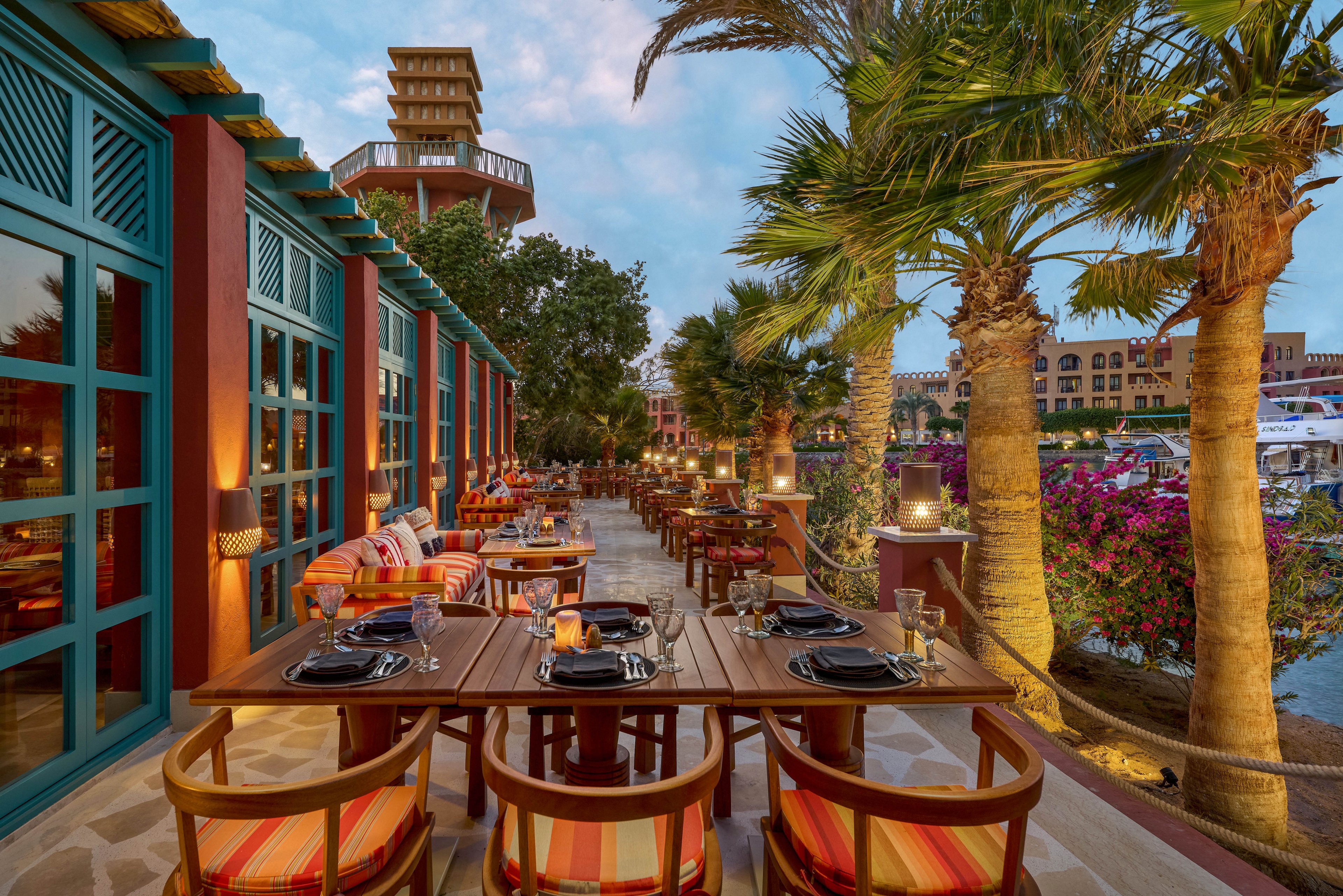 An inviting outdoor dining area set along a waterfront, featuring wooden tables and colorful striped chairs. The setting is enhanced by warm lighting, lush greenery, and a scenic view of boats and buildings in the background. The atmosphere is cozy and vibrant, perfect for an evening meal or gathering.