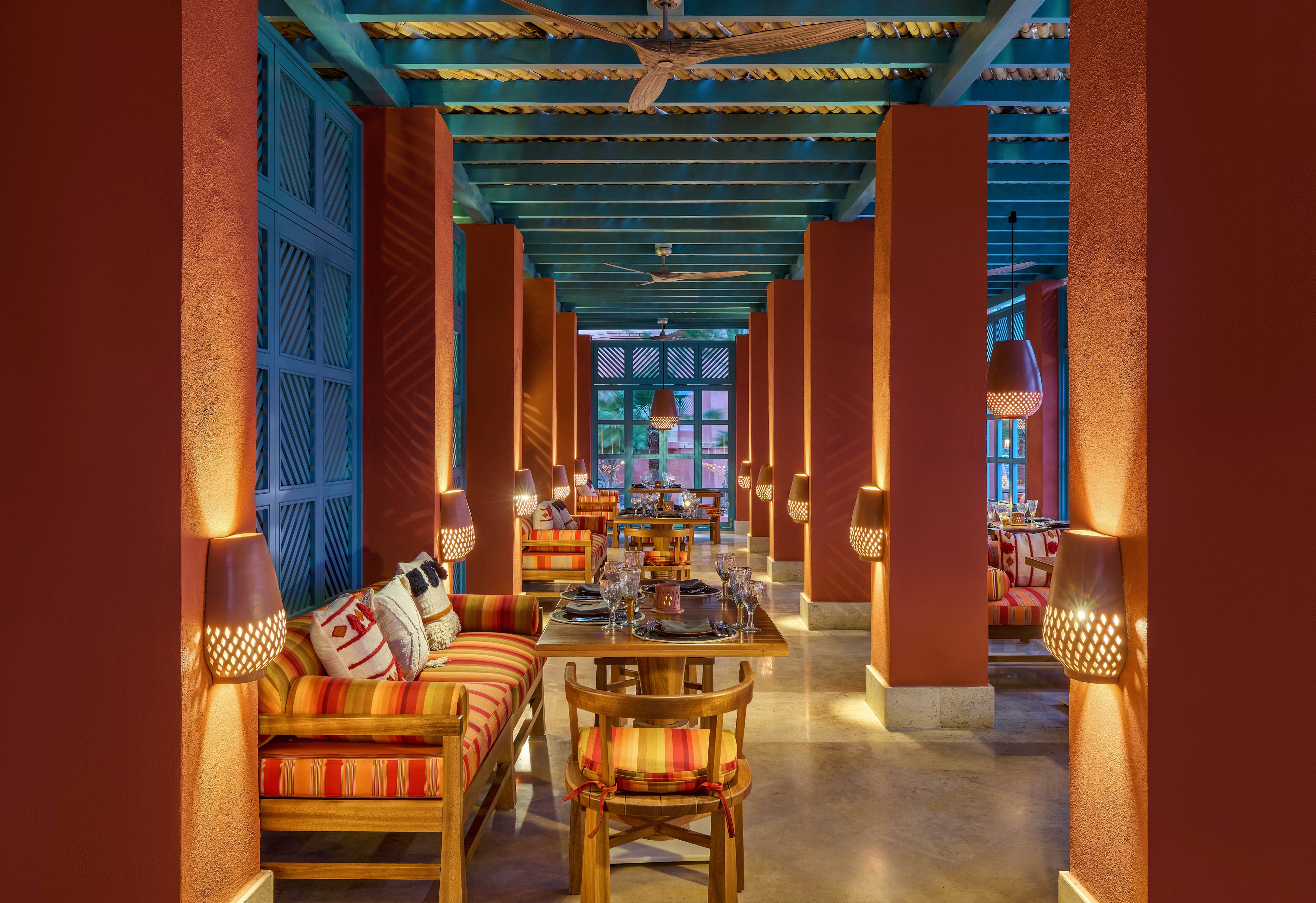 A warmly lit restaurant interior featuring vibrant red columns, striped cushions, and wooden furniture. The setting includes hanging pendant lights and a blue ceiling with wooden beams, creating a cozy and inviting atmosphere. The symmetrical arrangement of tables and chairs enhances the visual appeal.