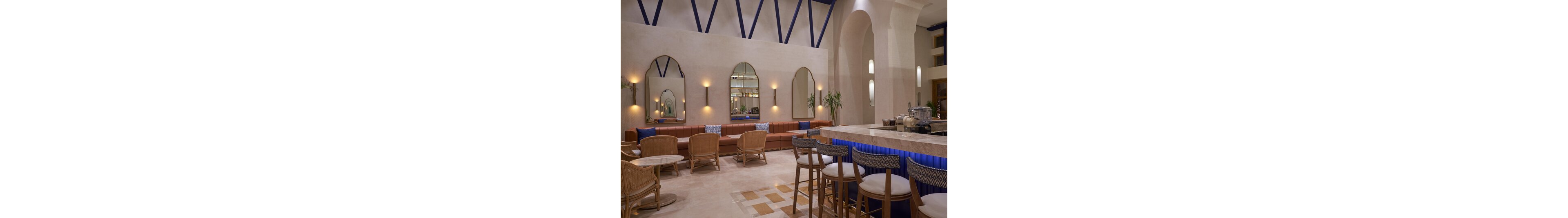 A contemporary hotel lounge bar features a long upholstered bench with small round tables and wicker chairs. A marble-topped bar with high stools and blue accent lighting stands in the foreground. Arched mirrors, wall sconces, and tall architectural arches create a refined, airy atmosphere. Neutral beige tones are contrasted with blue cushions and decor, giving the space a calm, inviting feel.