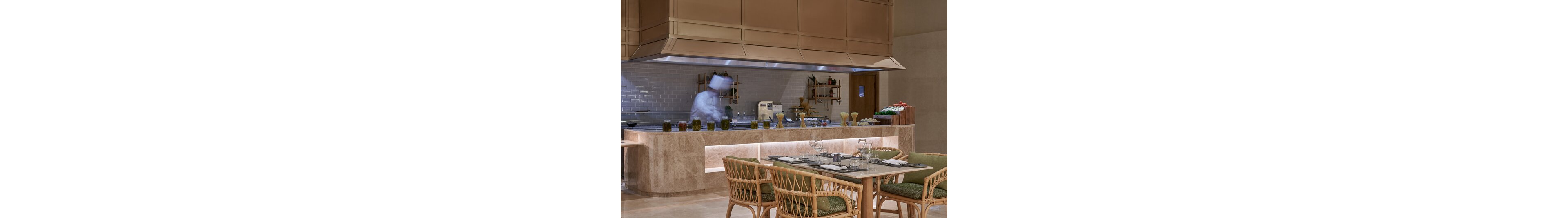 A modern open kitchen restaurant features a chef working behind a marble counter. The foreground shows a neatly set dining table with wicker chairs and green cushions. Shelves with jars, bottles, and kitchen tools line the tiled backsplash, adding visual interest. Warm lighting and neutral tones create an inviting, upscale dining atmosphere.