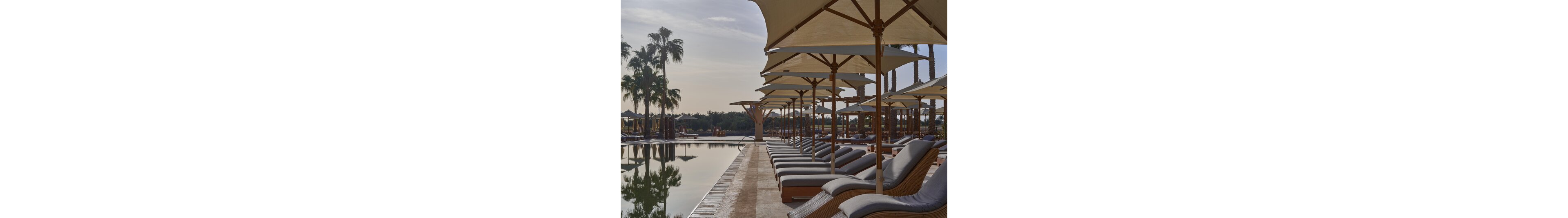 A row of cushioned wooden loungers lines the edge of a calm resort swimming pool. Large beige sun umbrellas provide shade along the pool deck, creating a uniform, orderly scene. Palm trees and landscaped greenery frame the background under a hazy sky, suggesting a warm, tranquil climate. The overall atmosphere feels quiet and relaxing, ideal for vacation or leisure time by the water.