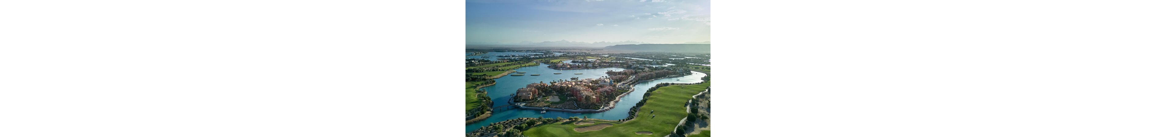 An aerial view captures a luxurious resort complex surrounded by winding blue waterways and lush greenery. The central island features clusters of colorful buildings, palm trees, and small bridges connecting different sections. A well-maintained golf course with fairways and greens stretches along the water’s edge. In the distance, multiple lagoons and low-lying developments extend toward a hazy mountain range under a clear sky. The overall scene conveys a tranquil, upscale vacation destination by the water.
