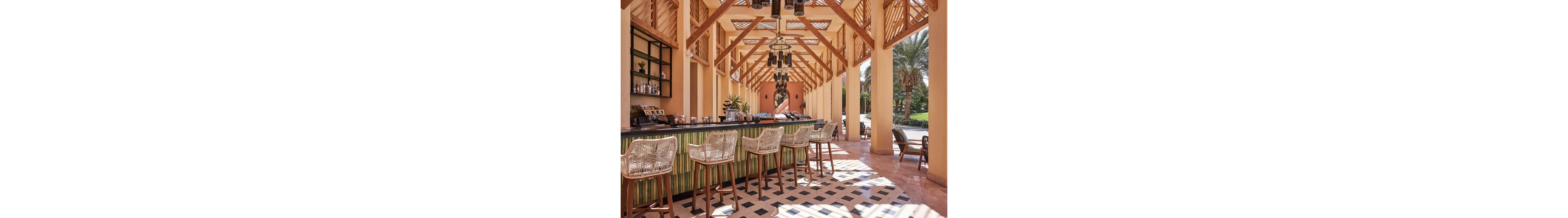 An open-air bar with a long counter and wicker bar stools sits under a high wooden pergola structure. The setting appears to be a tropical or resort environment with palm trees visible outside. Warm beige and terracotta tones dominate the architecture, complemented by a patterned black and white tiled floor. Hanging lantern-style lights and neatly arranged bottles on shelves add to the relaxed, upscale atmosphere.