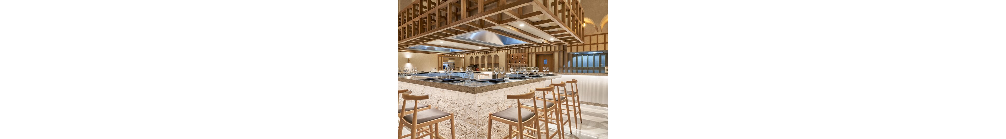 A contemporary restaurant interior features a central stone-clad bar with wooden stools arranged neatly around it. The open kitchen is framed by a geometric wooden ceiling structure and stainless steel hoods above the cooking area. Wine glasses and place settings are prepared on the countertop, suggesting a fine dining experience. Warm neutral lighting and beige tones create an inviting and sophisticated atmosphere.