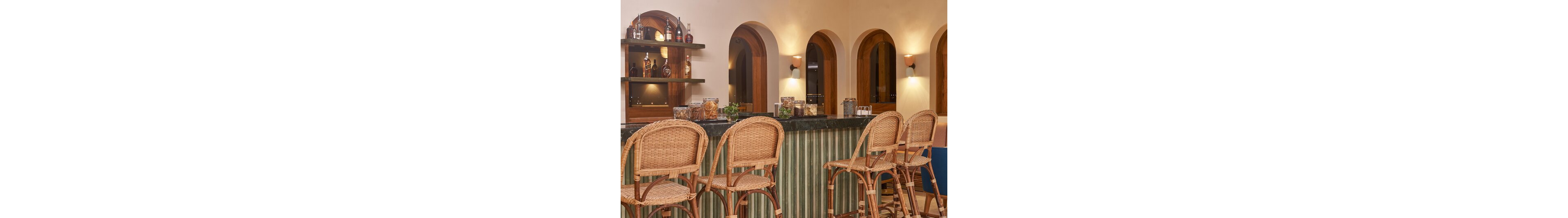 A warm, inviting bar interior features a dark stone countertop and several wicker bar stools neatly arranged along the front. Behind the bar, wooden shelves display assorted bottles and jars, adding visual interest and texture. Arched doorways and wall sconces create a soft, ambient glow throughout the space. The overall scene suggests a relaxed, intimate setting for drinks and conversation.