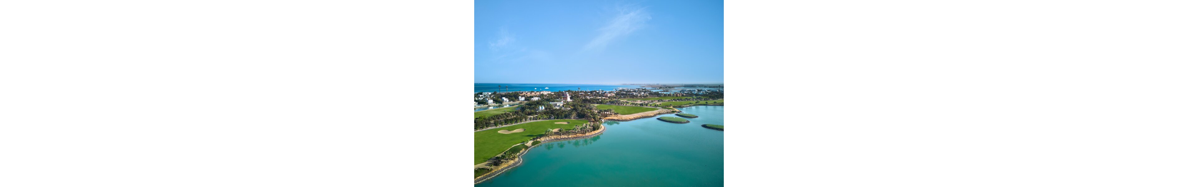 An aerial view showcases a lush golf course bordering a turquoise lagoon. Curved fairways, sand bunkers, and manicured greens are surrounded by palm trees and landscaped vegetation. Residential buildings and resort-style structures line the coastline in the background. The calm sea and clear blue sky create a serene, sunny coastal atmosphere.