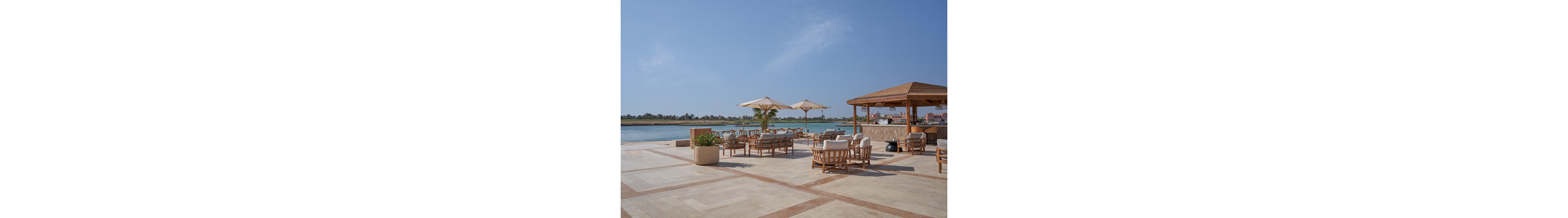 A spacious waterfront terrace features wooden lounge chairs and sofas arranged around low tables. Large cream-colored umbrellas provide shade over the seating area, while a wooden pergola structure houses a bar or service counter. The scene overlooks calm blue water with a distant shoreline lined with greenery and buildings. Clear skies and soft neutral tones create a relaxed, vacation-like atmosphere.