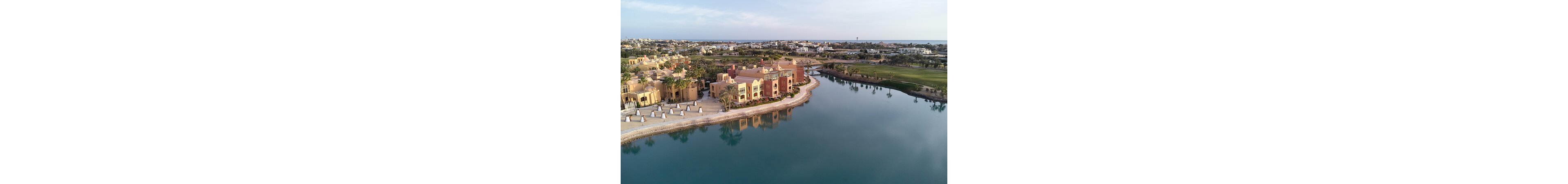 An aerial view shows a cluster of resort-style buildings beside a calm, blue lake. The architecture features warm earth tones and flat roofs, surrounded by palm trees and landscaped walkways. On the opposite shore, a manicured green area stretches toward the horizon under a clear sky. The wider background reveals additional low-rise structures and the distant coastline.