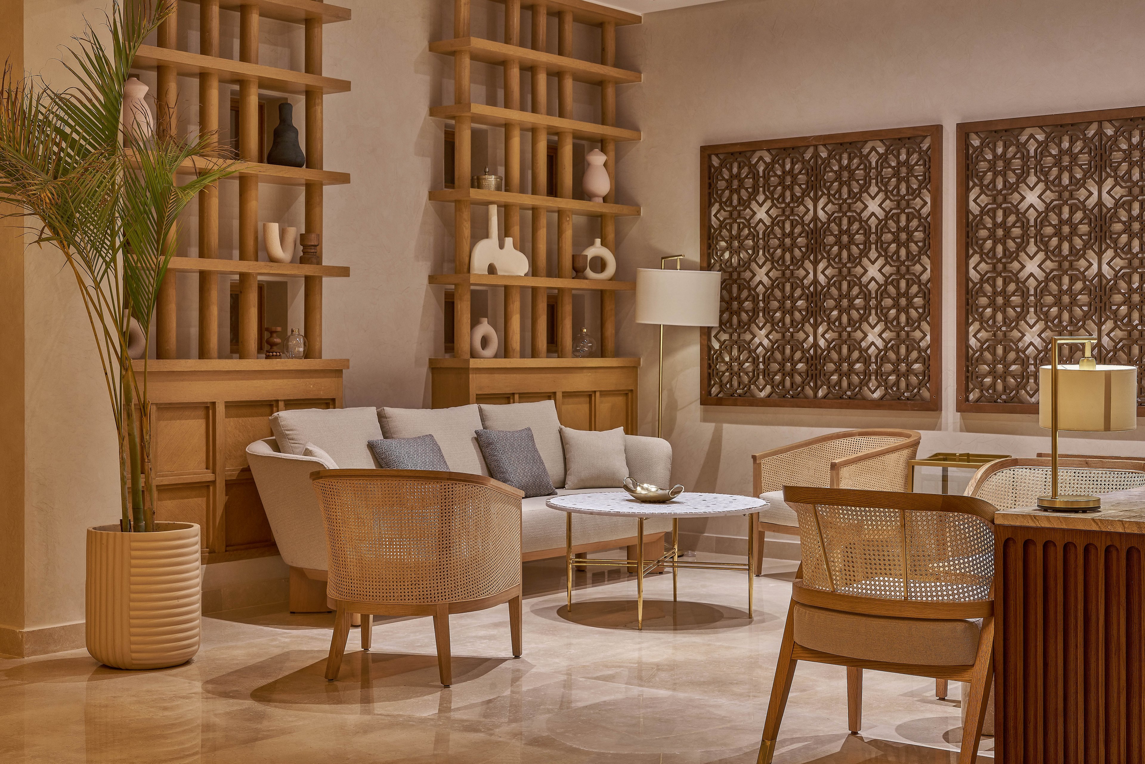 A stylish lounge area featuring neutral tones and minimalist decor. The space includes a white sofa with cushions, wooden chairs, and a round coffee table. Decorative shelves and intricate wall art enhance the ambiance, complemented by soft lighting and a potted plant.