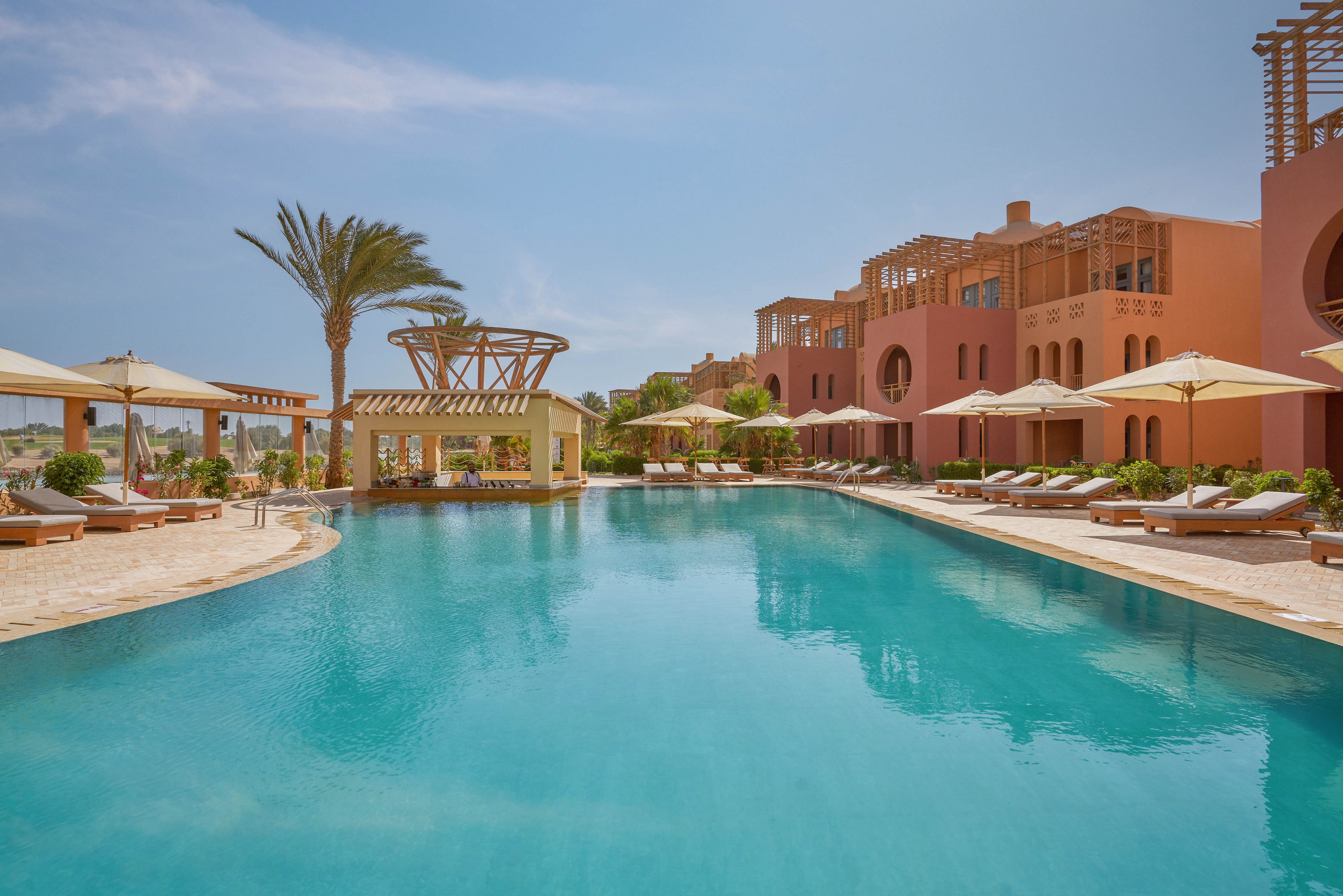 A serene outdoor pool surrounded by sun loungers and umbrellas at a luxury resort. The setting features vibrant terracotta-colored buildings and lush greenery under a clear blue sky. The tranquil atmosphere is ideal for relaxation and leisure.