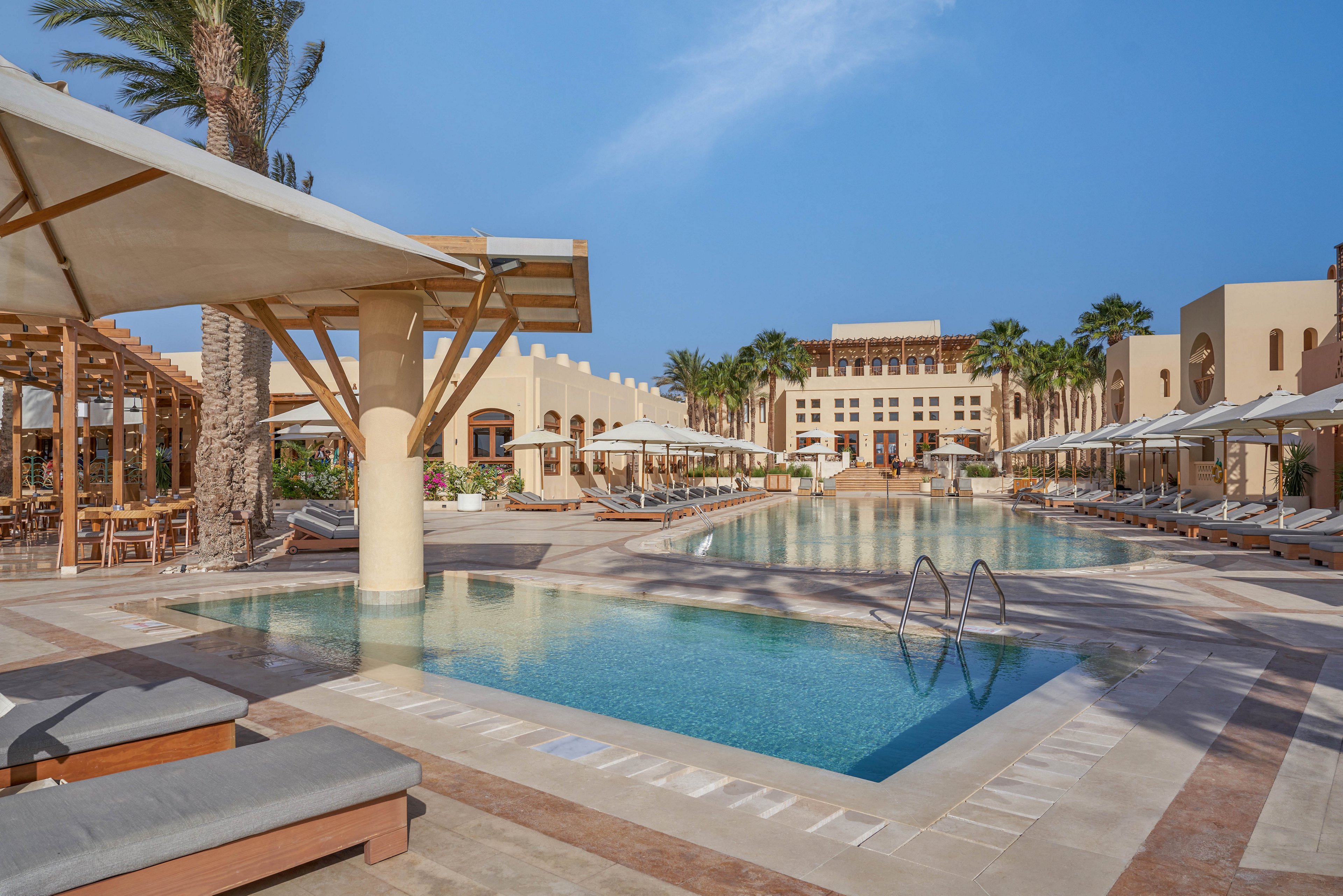 A serene luxury resort featuring a large swimming pool surrounded by sun loungers and umbrellas. The setting includes palm trees and modern architecture under a clear blue sky. The tranquil atmosphere is ideal for relaxation and leisure.