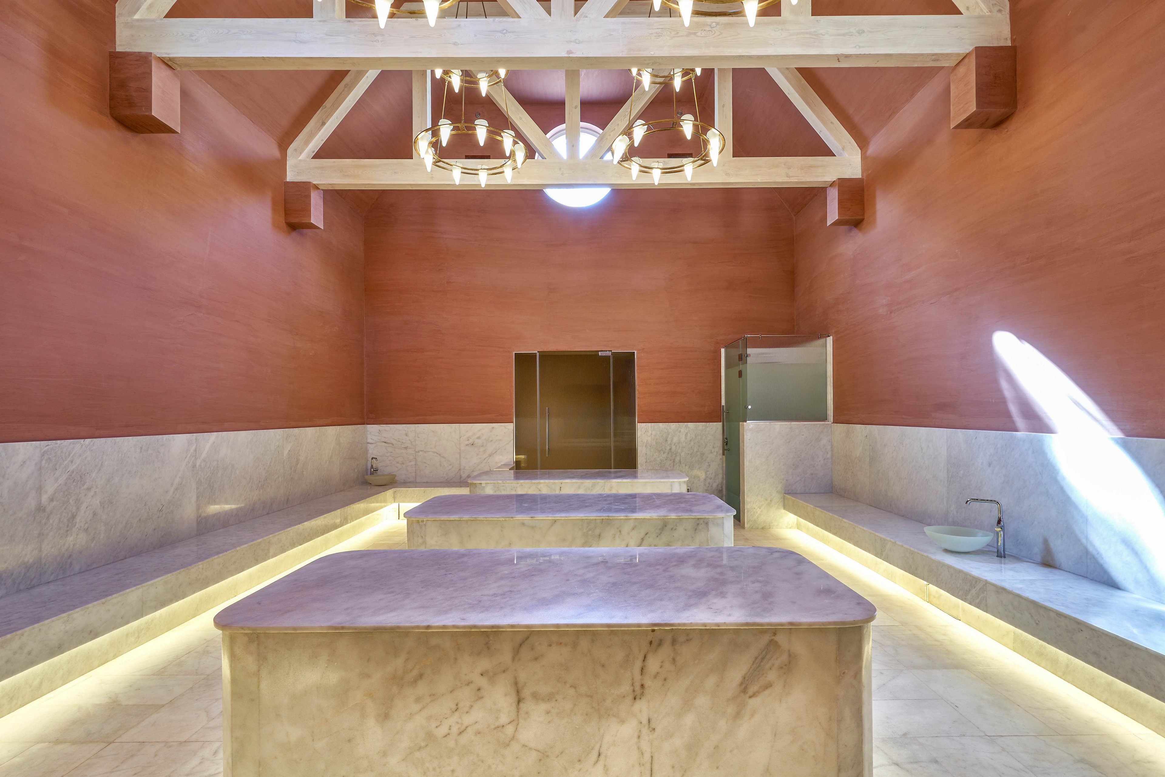 A spacious spa room featuring marble surfaces and elegant lighting. The room is illuminated by a large chandelier and natural light streaming through a circular window. The high wooden-beam ceiling adds to the luxurious ambiance, complemented by warm-toned walls and modern fixtures.