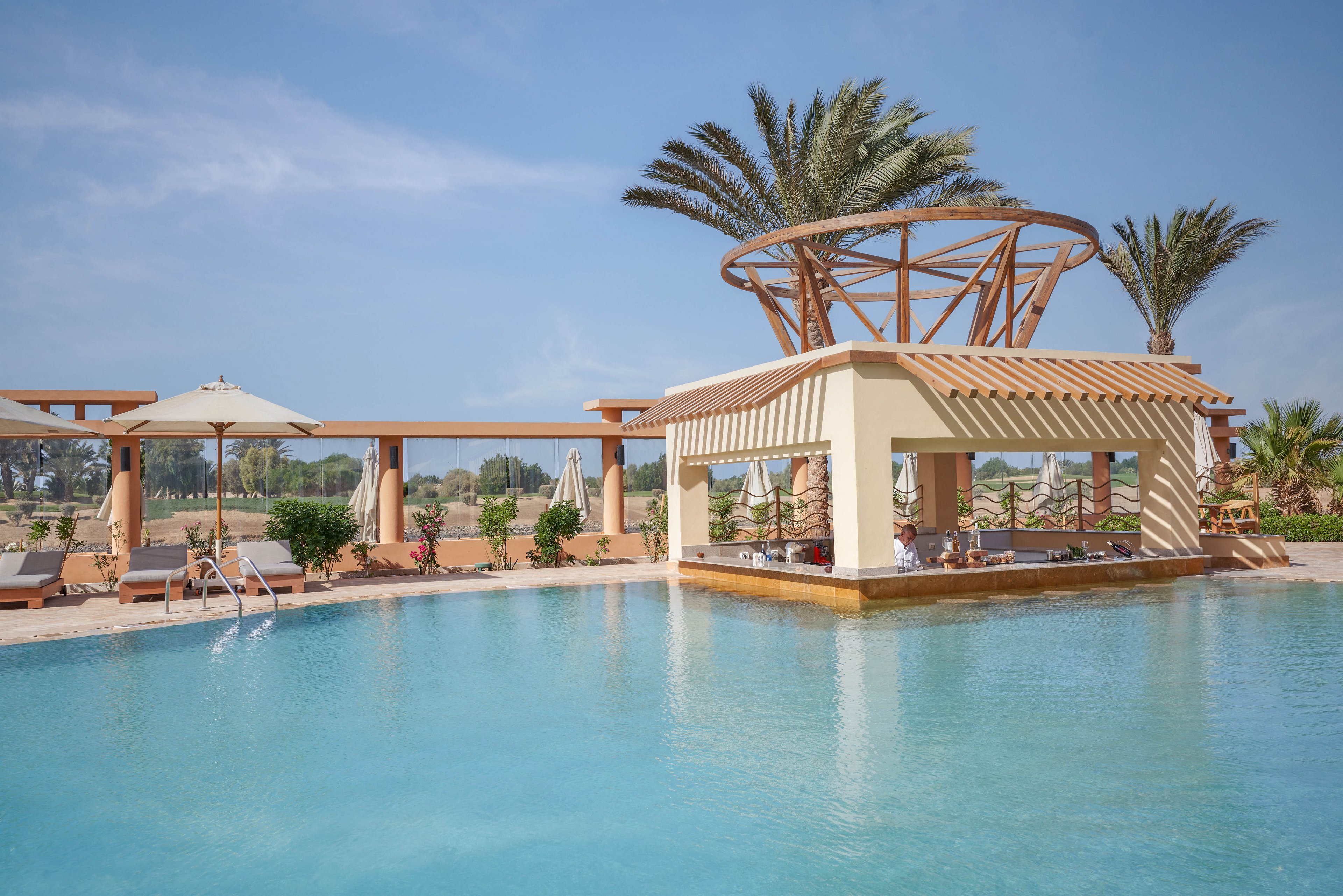 A serene outdoor poolside setting featuring a stylish pavilion with wooden accents and palm trees in the background. The clear blue water reflects the sunny sky, creating a tranquil atmosphere. Lounge chairs and umbrellas provide a relaxing space for guests.