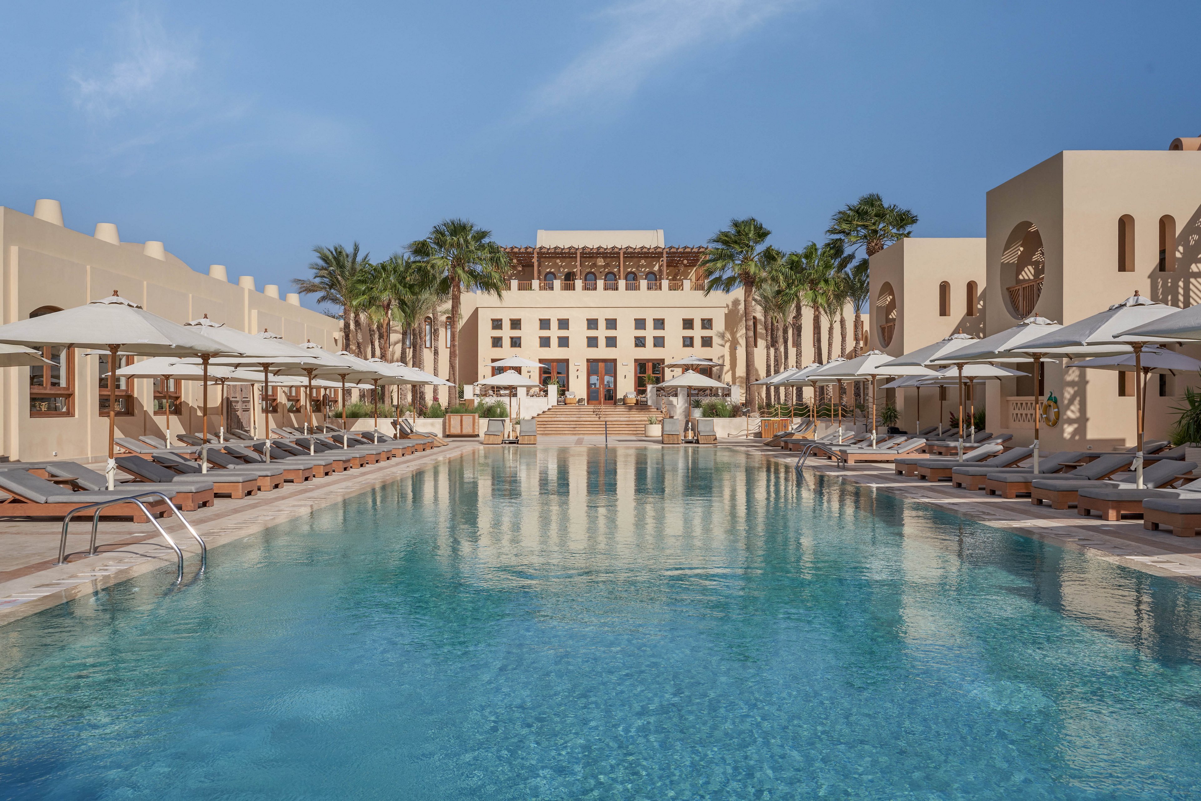 A serene luxury resort featuring a large swimming pool surrounded by sun loungers and umbrellas. The setting includes palm trees and beige architectural structures under a clear blue sky. The symmetrical design and tranquil atmosphere evoke relaxation and elegance.